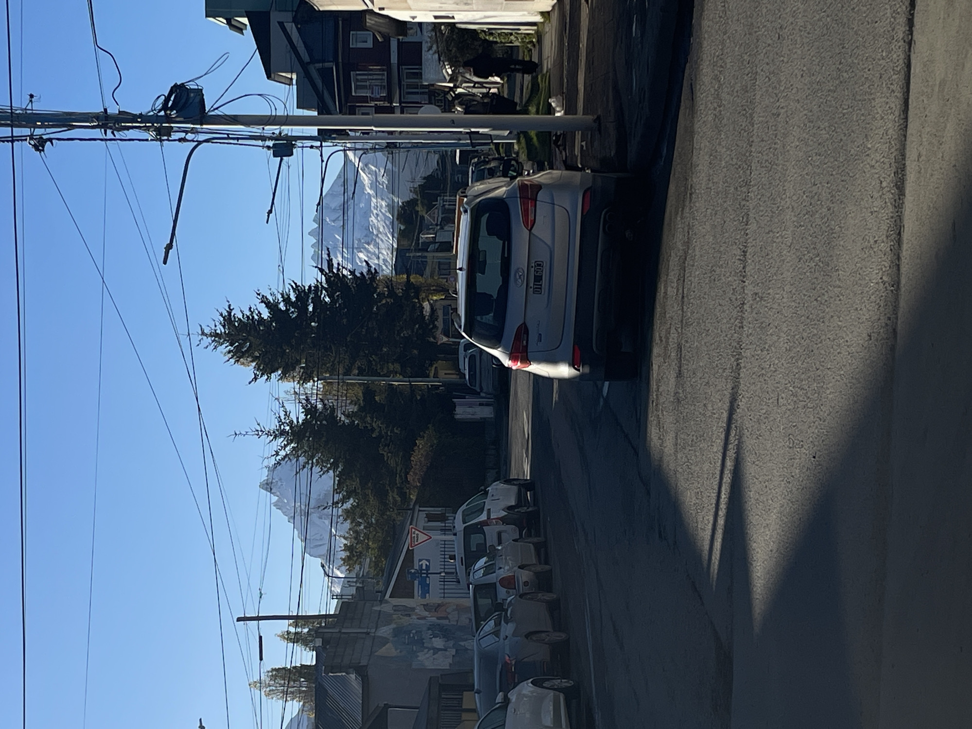 The image shows a street scene in Ushuaia, Argentina, with a line of parked cars along the right side of the road. A silver Hyundai SUV is prominently parked with the license plate "OIL 603" visible. Trees and snow-capped mountains are visible in the background. Overhead power lines run across the sky. Buildings line the street, including structures with dark-colored roofs. A person walks in the distance. Shadows are cast on the right side of the road, suggesting bright sunlight.