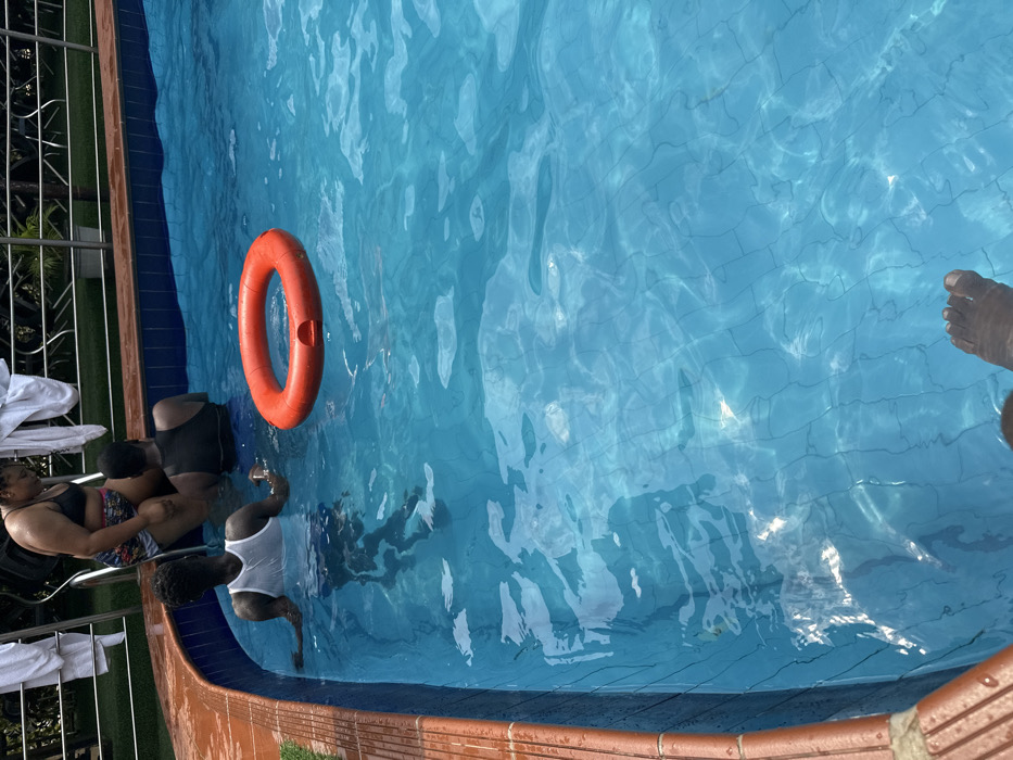 This image depicts a daytime scene at a swimming pool in Aba, Nigeria. Four individuals are present. One individual, wearing a dark bikini top and patterned shorts, is partially submerged near a metallic pool ladder at the pool's edge. Behind this individual, white towels are draped over a silver railing. Another individual, seen from behind and wearing a white tank top, is partially submerged, resting forearms on the pool's brown-tiled edge. A third individual, also seen from behind and wearing a black tank top, is submerged deeper within the pool. An orange ring buoy floats in the clear blue water. The feet of a fourth individual are visible in the foreground, submerged in the water. The pool features blue mosaic tiles on its interior walls and bottom. Green foliage and a metal fence are visible in the background beyond the pool's edge.