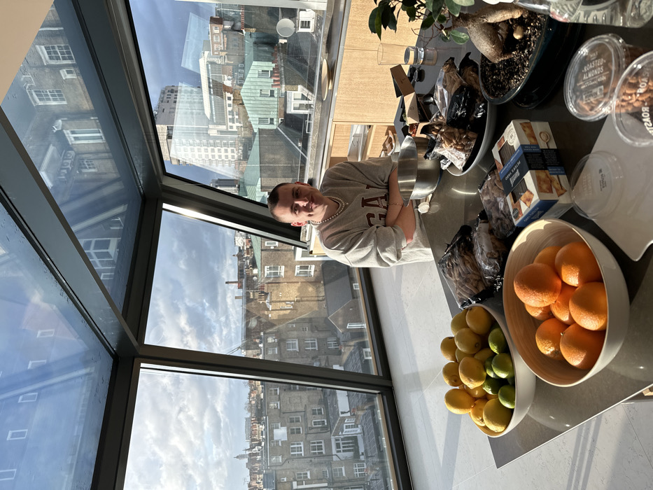 The image, oriented sideways, captures a bright daytime scene inside a modern apartment or building, offering a panoramic view of a city. Through large, framed windows, numerous older buildings with brick facades and varied rooflines are visible under a partly cloudy sky, consistent with the architecture of London.
In the foreground, a young man, likely in his 20s or 30s, is seated or standing behind a reflective grey countertop. He has short hair and is wearing a light grey sweatshirt with the letters "UCLA" (or similar college branding) visible on the front. He sports a pearl necklace and a bracelet, has his arms crossed, and is smiling directly at the camera, conveying a relaxed and happy demeanor.
The countertop is laden with various items. Two large, light-colored bowls are filled with fresh fruit: one with oranges and another with lemons and limes. Nearby, several packages of food or snacks are arranged. These include two clear plastic containers clearly labeled "ROASTED ALMONDS" and a cardboard box partially showing "PETER'S Y... BISCUITS," suggesting crackers or similar baked goods. Several other bags, appearing to contain nuts or dried snacks, are also present. A large silver bowl or pot and a potted plant with a thick trunk and sparse foliage in a black pot are also on the surface. The windows and countertop exhibit reflections of the interior space and the exterior sky.