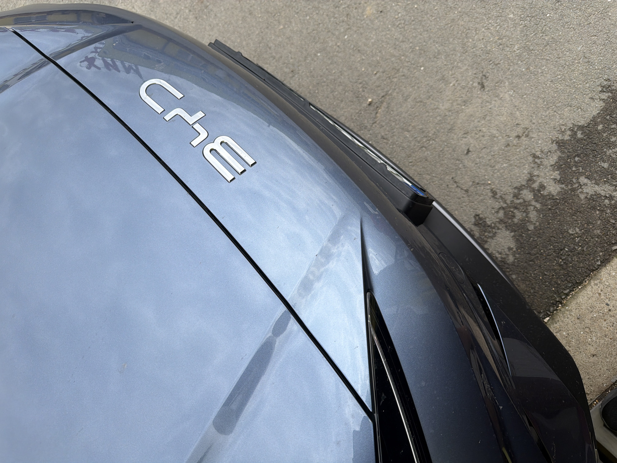 The image shows the front of a car, likely a BYD, with its logo prominently displayed. The car is a metallic blue-grey color. The logo, composed of chrome-plated letters, reads "BYD". A black line separates the hood of the car from the bumper area. The car is parked on a paved surface, which appears to be asphalt or concrete, with some patches of wear and discoloration. The environment suggests an outdoor urban or suburban setting. There are no people visible in the image. The focus is on the car's front emblem and hood. The location of Dolno Ablanovo, Bulgaria is not discernible from the visual cues in the photograph.