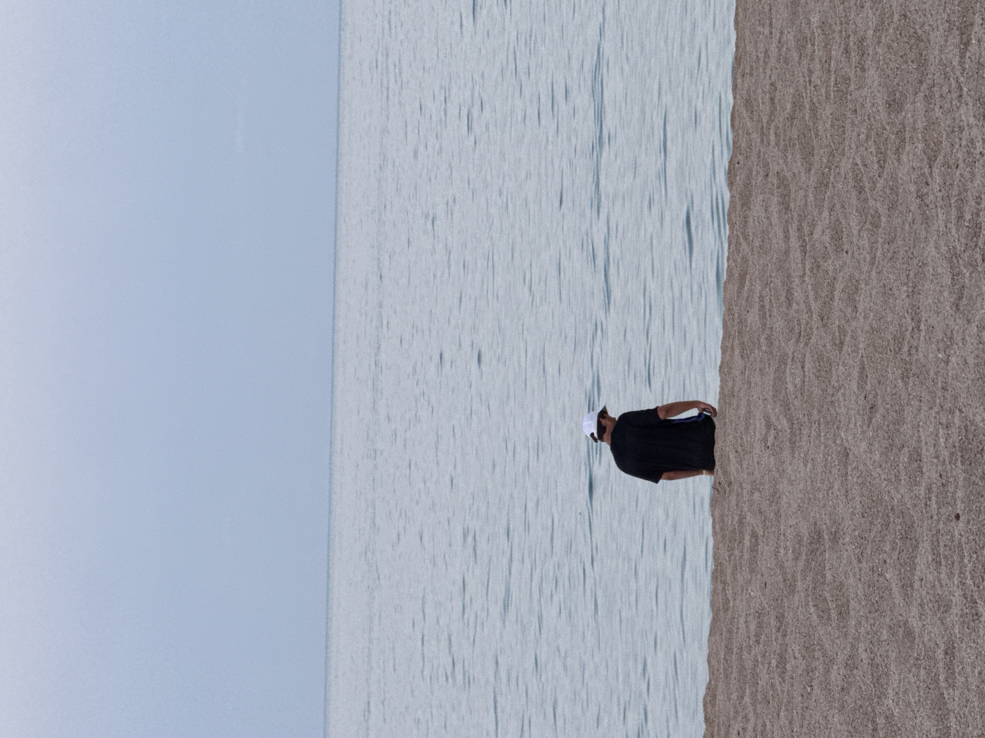 A lone person, viewed from behind, stands at the edge of the water on a beach. The individual is wearing a dark t-shirt and a white baseball cap. They are facing the ocean, with their head bowed. The sand of the beach is a light brown color and appears to be composed of small pebbles. The water is a pale blue-gray and calm, with gentle ripples. The sky above is overcast and a similar pale blue-gray color, meeting the horizon with a distinct line. There are no other people or objects visible in the frame. The scene suggests a quiet, contemplative moment.