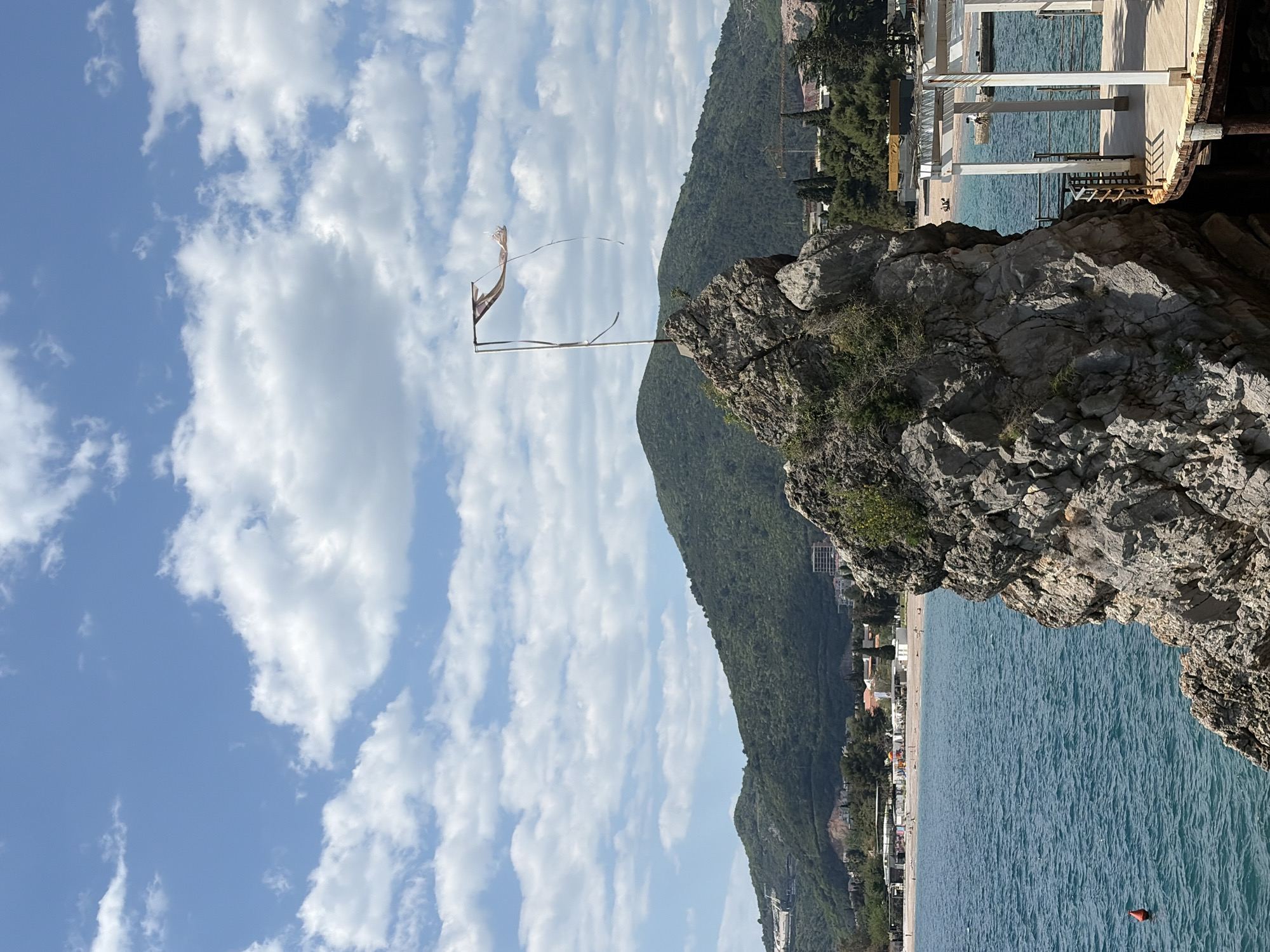 The image captures a scene in Budva, Montenegro, featuring a rocky cliff overlooking the Adriatic Sea. A tattered flag is attached to a makeshift flagpole on the cliff's edge. Behind the cliff, a green mountain slopes upwards, dotted with trees. The coastline is visible, with a beach and buildings running along the shore. The sea is blue, and a small red buoy floats on its surface. The sky is blue with scattered white clouds. A structure with white pillars is perched on the edge of the cliff.