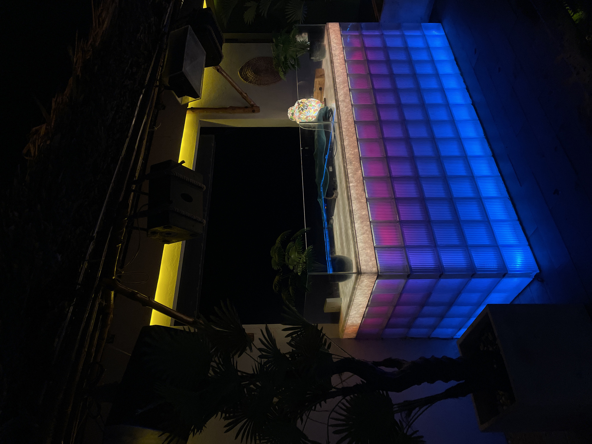 The image captures a vibrant and modern architectural facade at night in Phu Thuy Ward, Vietnam. The central feature is a large section of the building made of glass blocks, illuminated from within to display a striking gradient of colors. On the left, these blocks glow with pink and magenta hues, gradually transitioning to bright blue on the right, creating a dynamic light display.
To the left of the glass block section, a vertical strip of warm yellow light illuminates a textured wall. Below this, dark, rectangular speaker-like boxes are mounted, suggesting a public or entertainment space. Further left, dark silhouettes of tropical plants, possibly palm fronds, are visible against the deep night sky, and what appears to be a ladder or series of bamboo poles leans against the wall.
Above a reflective surface in the mid-ground, a unique, colorful mosaic-like light fixture or sculpture casts its intricate patterns, which are mirrored in the surface below. Additional dark foliage from large plants frames the lower right and left corners of the scene.
No human activity or individuals are present in the image. The scene is static, highlighting the architectural design and its sophisticated lighting. There is no visible text. The overall atmosphere is modern, nocturnal, and inviting, emphasizing the interplay of light, color, and texture.