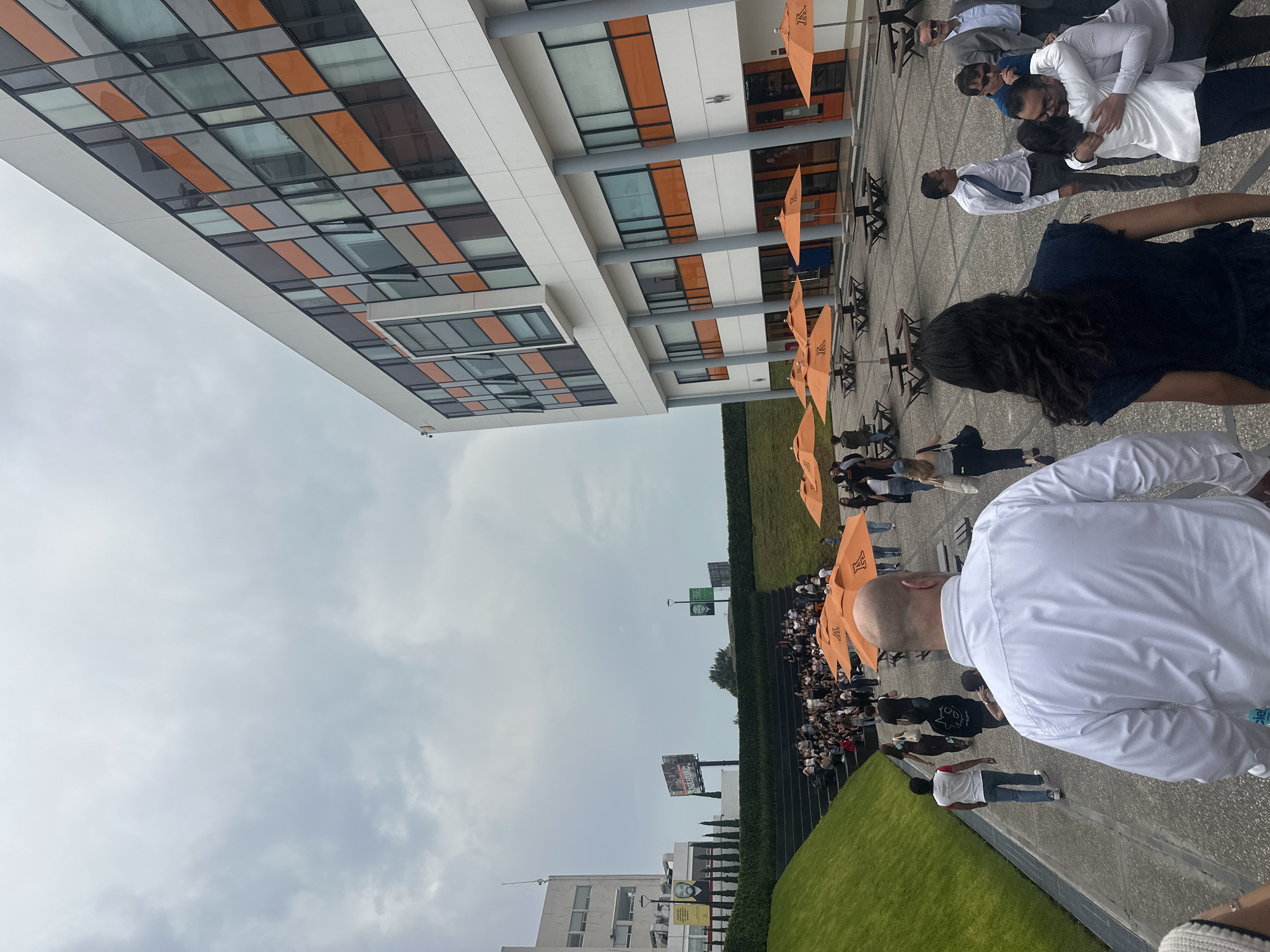 The image shows an outdoor scene at what appears to be a graduation ceremony, possibly in the city of San Bernardino Tlaxcalancingo, Mexico. A large group of people, many in white shirts, are seated in rows, likely graduates. Orange umbrellas provide shade. The background includes a modern building with large, rectangular windows and a white exterior. The sky is overcast. The foreground features a walkway where people in formal attire are congregated. Some individuals are embracing and interacting. A section of green lawn and a high fence run parallel to the seating area. A billboard is visible in the distance. The image is slightly angled.