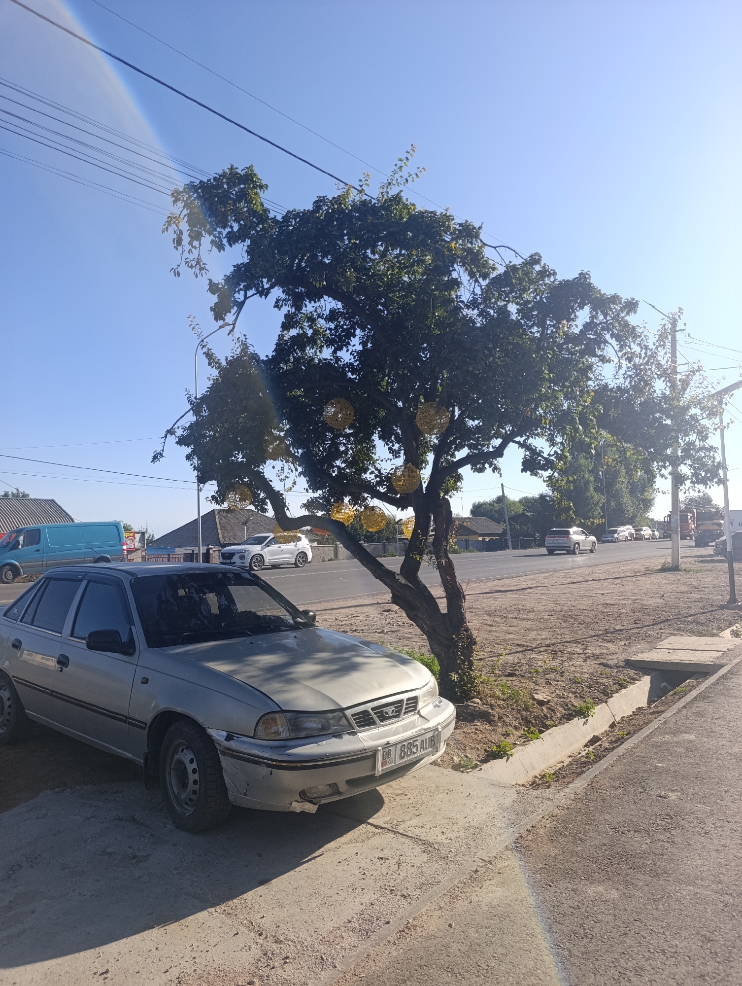In Bosteri, Kyrgyzstan, a silver Daewoo Nexia sedan is parked on a dirt shoulder next to a paved road. The car is angled towards the viewer, with its front bumper and driver's side visible. A large, leafy tree with decorative yellow spheres hanging from its branches is situated behind the car. The tree casts a shadow on the car and the ground. In the background, a blue van is parked on the left side of the road, and several other vehicles, including a white SUV and a truck, are seen driving on the road. The sky is clear and blue, with sunlight casting a rainbow-like lens flare on the lower right side of the image. The environment appears to be a semi-urban or rural setting, with buildings and vegetation visible in the distance.