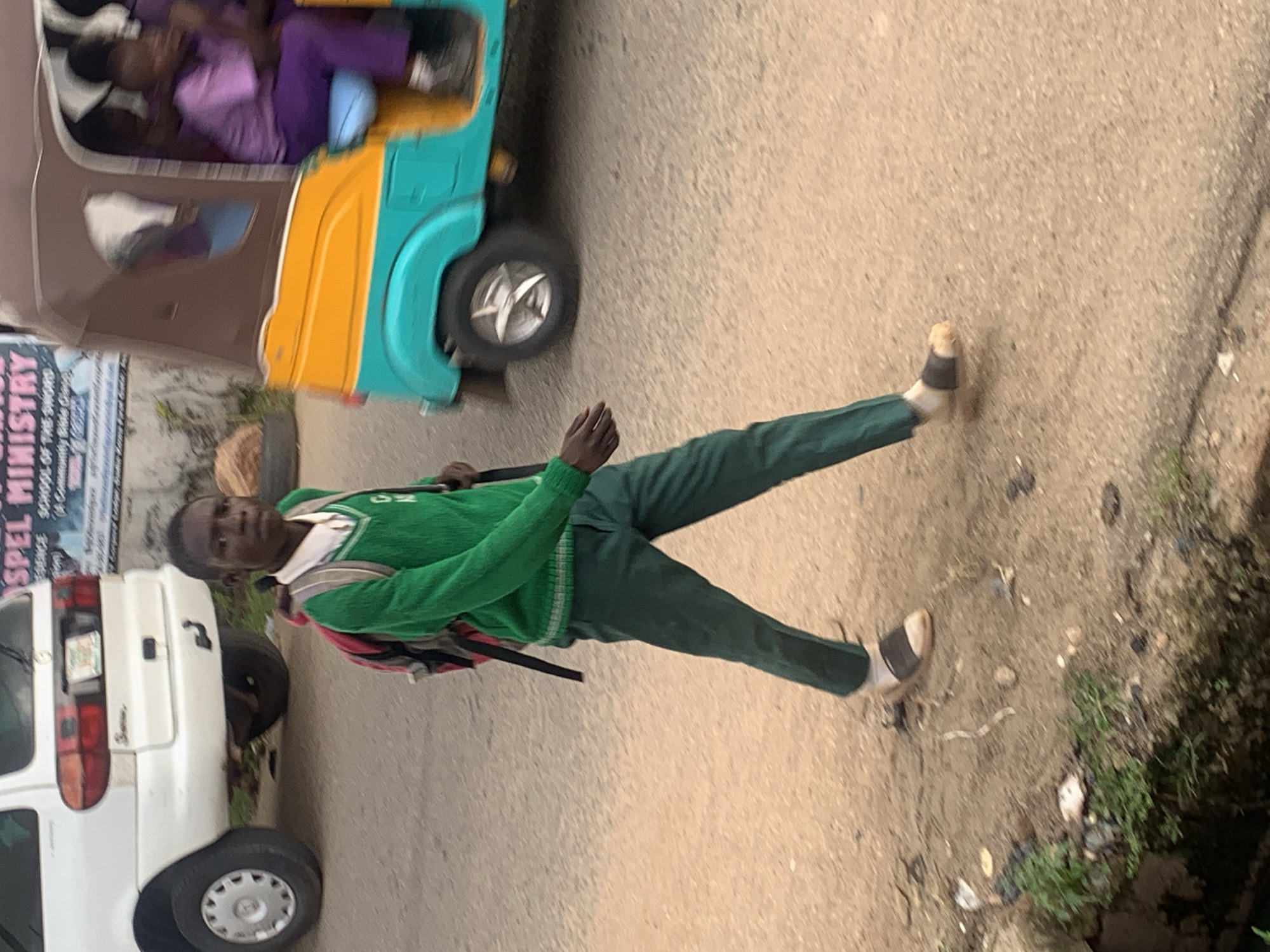 A young boy wearing a green sweater and green pants stands on a dirt road. He has a backpack on his shoulders. To his left is a white van. Behind the van is a colorful auto-rickshaw with people inside. The auto-rickshaw is turquoise and yellow. There is some text on a poster behind the white van. The text appears to be in English and mentions "GOSPEL MINISTRY". The scene appears to be outdoors, and the lighting suggests it could be daytime. The ground is rough and uneven, with some vegetation visible on the right side of the image.