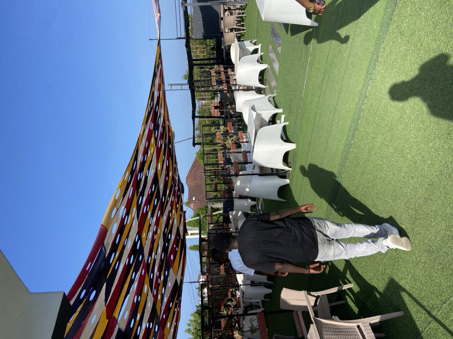 This image captures an outdoor leisure or event space under a clear, bright blue sky, likely during midday or early afternoon due to the strong sunlight and distinct shadows. The ground is covered with artificial green turf.
In the foreground, a person is seen from behind, wearing a black short-sleeved shirt, light grey pants, and white sneakers, standing on the turf. Another person's forearm and hand, holding a brown object, are visible on the right. Long, pronounced shadows of individuals and objects stretch across the green surface.
The midground features rows of modern, white, possibly plastic or rattan-style chairs, neatly arranged on the turf. Beyond these, additional darker, wicker-style chairs and tables are visible. A black metal railing or fence runs horizontally across the midground, with small potted plants placed on a ledge behind it.
A prominent feature in the left and upper parts of the frame is a large, colorful fabric sunshade or awning. It displays a vibrant geometric pattern of red, yellow, black, and light-colored stripes and shapes, interspersed with numerous circular cutouts that allow light to filter through. The background shows glimpses of a brown-tiled roof and some distant greenery, hinting at surrounding structures or natural elements.
The setting appears to be a rooftop lounge or an elevated outdoor venue, designed for comfort and social gatherings, consistent with a sunny location such as Dar es Salaam, Tanzania. No visible text is present in the image.