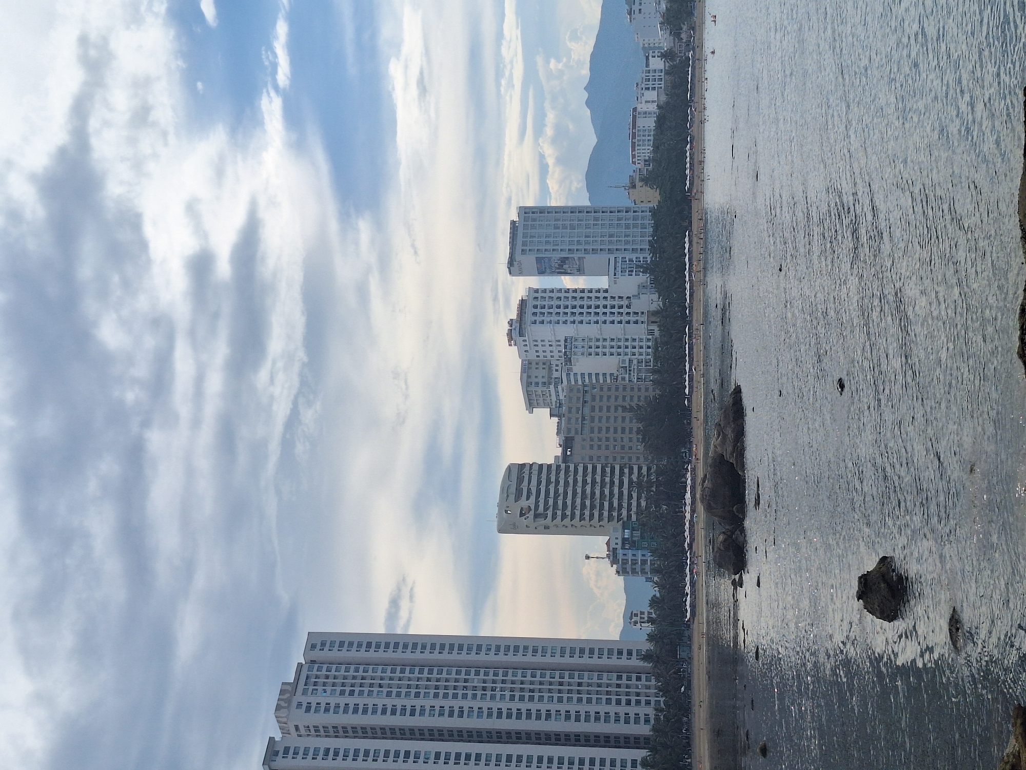 The image depicts a coastal city scene, likely in Nha Trang, Vietnam, characterized by a beach, ocean, and urban development. In the foreground, the ocean occupies a significant portion of the frame, with gentle ripples on its surface reflecting the cloudy sky. Several large rocks are visible, some partially submerged in the water, indicating a rocky coastline or a shallow bay.
The middle ground showcases a sandy beach lined with a dense arrangement of palm trees. Numerous white umbrellas are set up on the beach, suggesting it is a public area for recreation and relaxation. A few small figures of people can be discerned on the beach, engaged in various activities like walking or sunbathing.
Dominating the background is a skyline of modern, multi-story buildings. The most prominent structure on the far left is a very tall, white high-rise apartment building with a uniform facade of many windows. To its right, a series of other hotels and residential buildings of varying architectural styles and heights are visible. Some buildings feature unique designs, such as one with curved balconies. In the distance, beyond the city buildings, a range of mountains can be seen, their peaks softened by the haze and the overcast sky. The sky itself is a mix of light and dark gray clouds, with patches of blue sky peeking through, indicating a possibly humid or transitional weather condition. The overall impression is one of a vibrant coastal city that blends natural beauty with urban infrastructure.