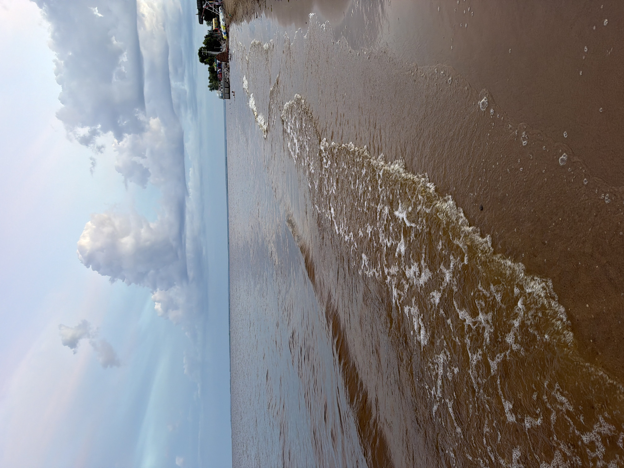 The image depicts a calm beach scene in Laishevo, Russia, with gentle waves lapping the shore. The water is a murky brown, suggesting it may be a river or a lake rather than the open sea. The sky above is a mix of pale blue and grey, with fluffy white clouds scattered across it. In the distance, along the shoreline, there are some trees and structures, including what appears to be a pier or a small dock with buildings and a yellow and white vehicle parked on the sand. A few people can be seen in the water, swimming or wading. The sand on the beach is a light brown color, and there are some scattered bubbles and ripples on the wet sand where the waves have receded. The overall impression is one of a peaceful, somewhat overcast day at the beach.