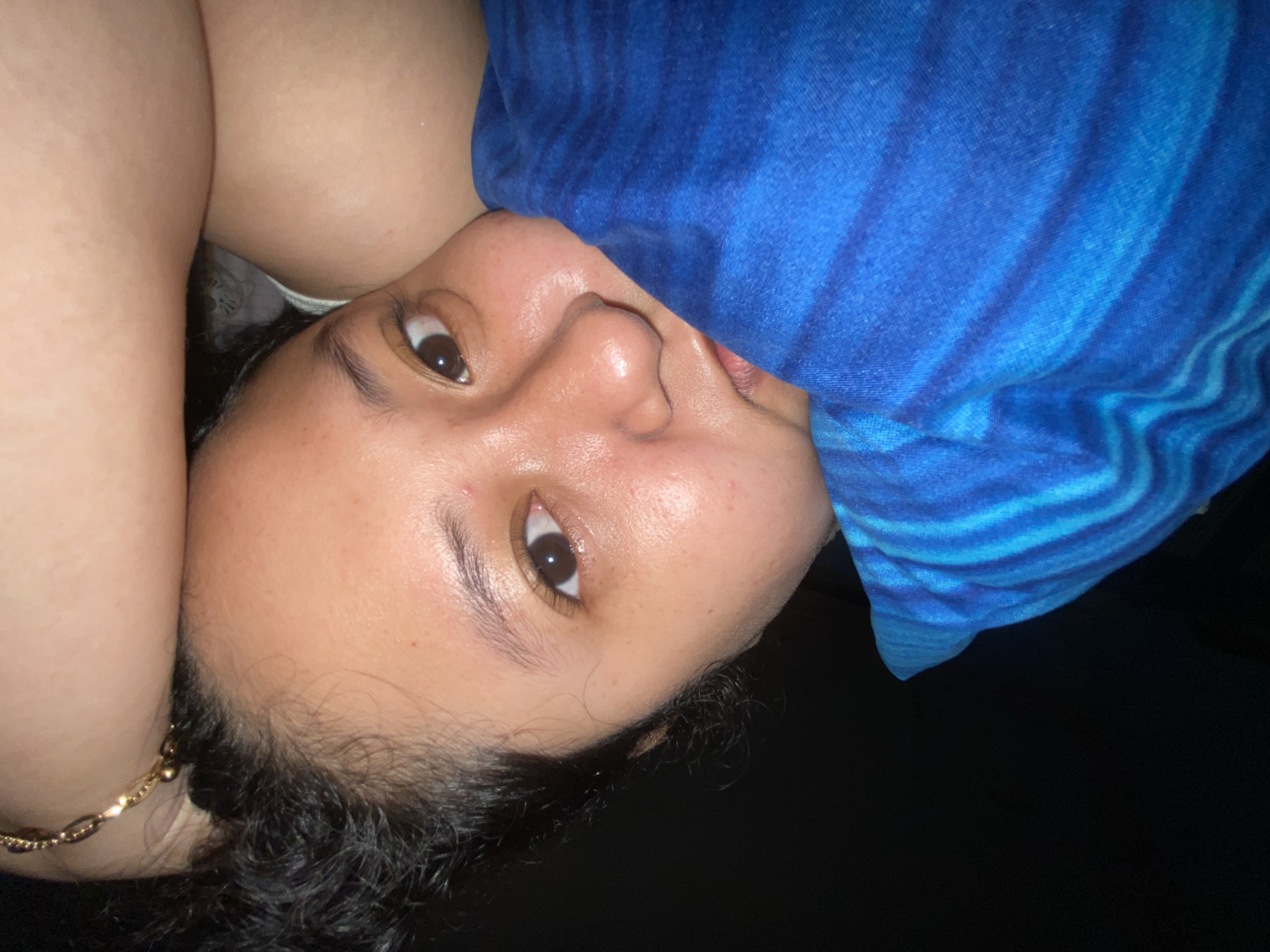 A close-up, overhead shot shows a person lying down with their head resting on their arm, covered by a blue striped blanket. The person's face is partially visible, with one eye looking directly at the camera. Their hair is dark and curly, and they are wearing a gold bracelet on their wrist. The background is dark, suggesting an indoor setting at night. No specific location in Gelam, Indonesia is discernible from the image.