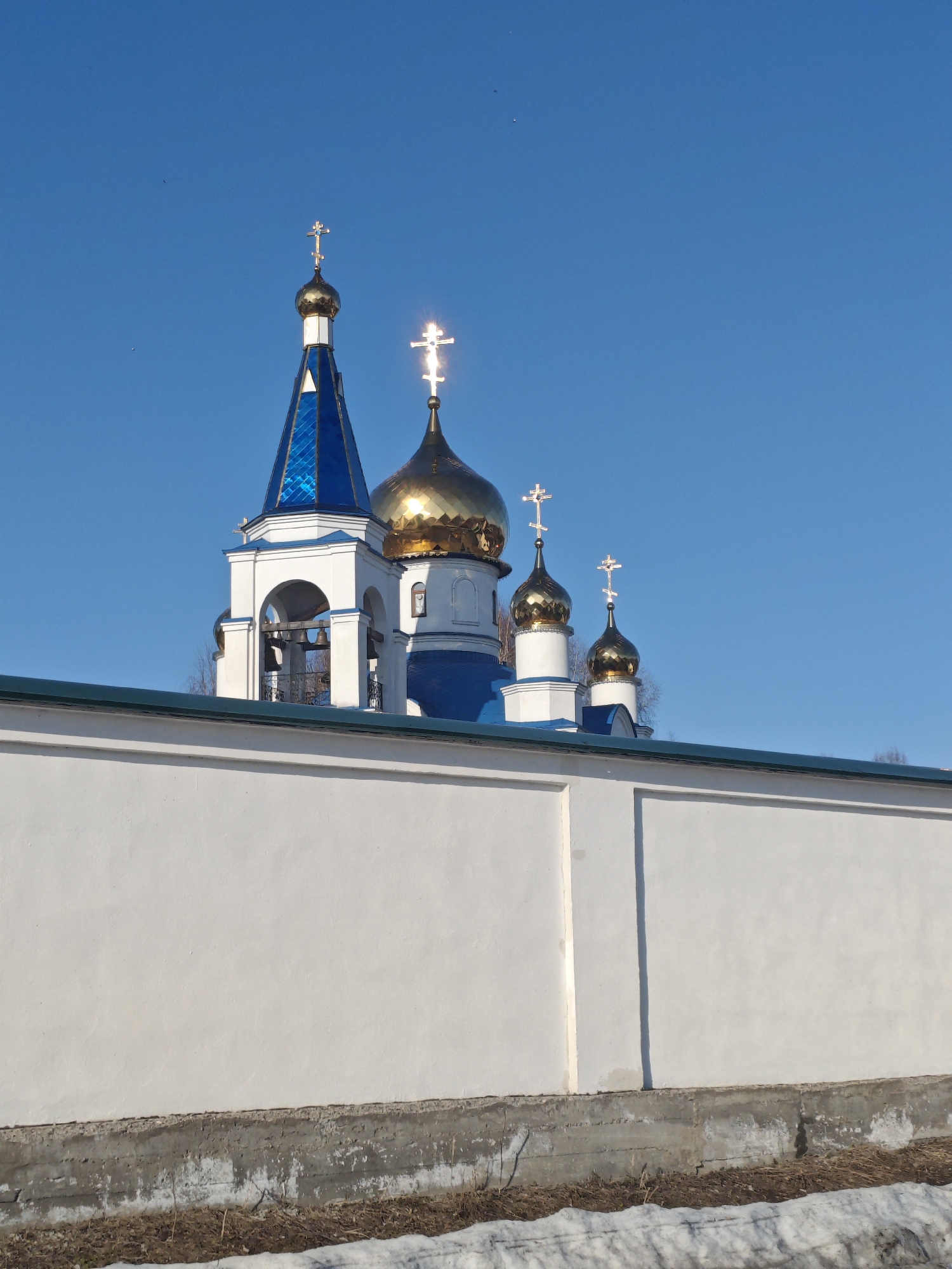 The image displays an Orthodox Christian church or monastery complex partially visible above a high wall, under a clear blue sky. In the upper portion of the frame, three white structures with blue elements and golden domes are prominent. From left to right, a tall bell tower features a pointed, faceted blue roof culminating in a silver-colored dome topped with an Orthodox cross. Below this, arched openings reveal multiple bells. Adjacent to the bell tower, a large golden dome with an Orthodox cross gleams brightly in direct sunlight. To its right, two smaller golden domes, each topped with an Orthodox cross, are visible, with the rightmost cross also reflecting intense sunlight. The structures are primarily white with blue roofs and golden cupolas.
The lower half of the image is dominated by a plain, tall white perimeter wall, capped with a dark green coping. Vertical architectural details or shadows divide the wall into sections. Below the white wall, a concrete base is visible, showing signs of wear and some discoloration. At ground level, a strip of dry, brown vegetation is present, with patches of snow evident on the far right. The overall environment suggests a cold climate or late winter/early spring conditions. The sky is uniformly clear and deep blue. No people or overt actions are depicted. Several small dark specks, likely birds, are discernable against the blue sky. The location is an unidentified city in an unknown region.