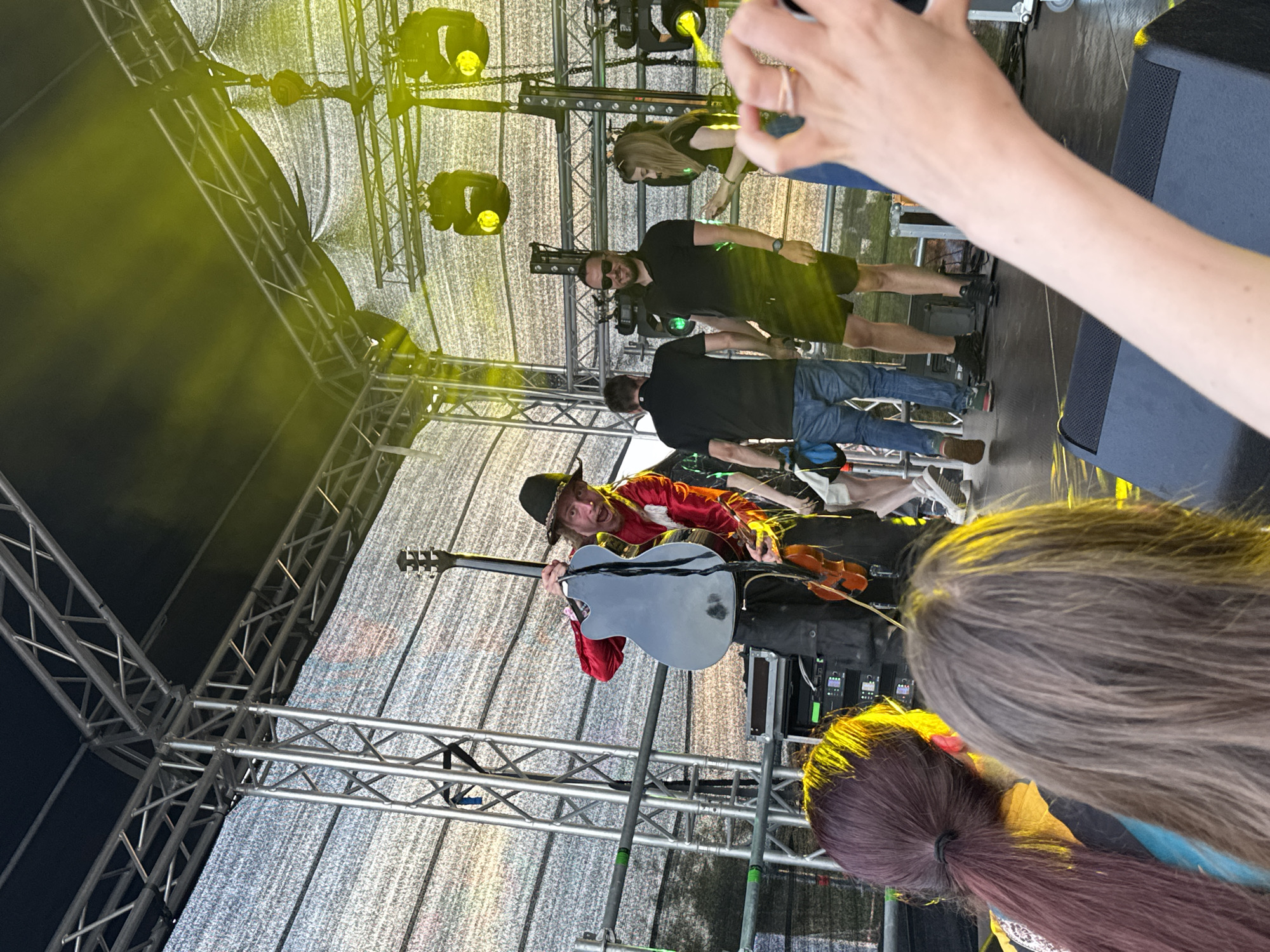 On a brightly lit outdoor stage, a performer in a red shirt and black hat holds a dark gray guitar while a violin is visible below. The stage is constructed from silver metal trusses and draped with white mesh fabric, illuminated by yellow stage lights. Several people are on the stage, including men in black shirts and shorts, and a woman in a black top and jeans. In the foreground, two audience members with their backs to the camera, one with dark red hair in a ponytail and the other with gray hair, are visible. The scene appears to be a live music performance in Tallinn, Estonia, possibly at the Tallinn Song Festival Grounds.