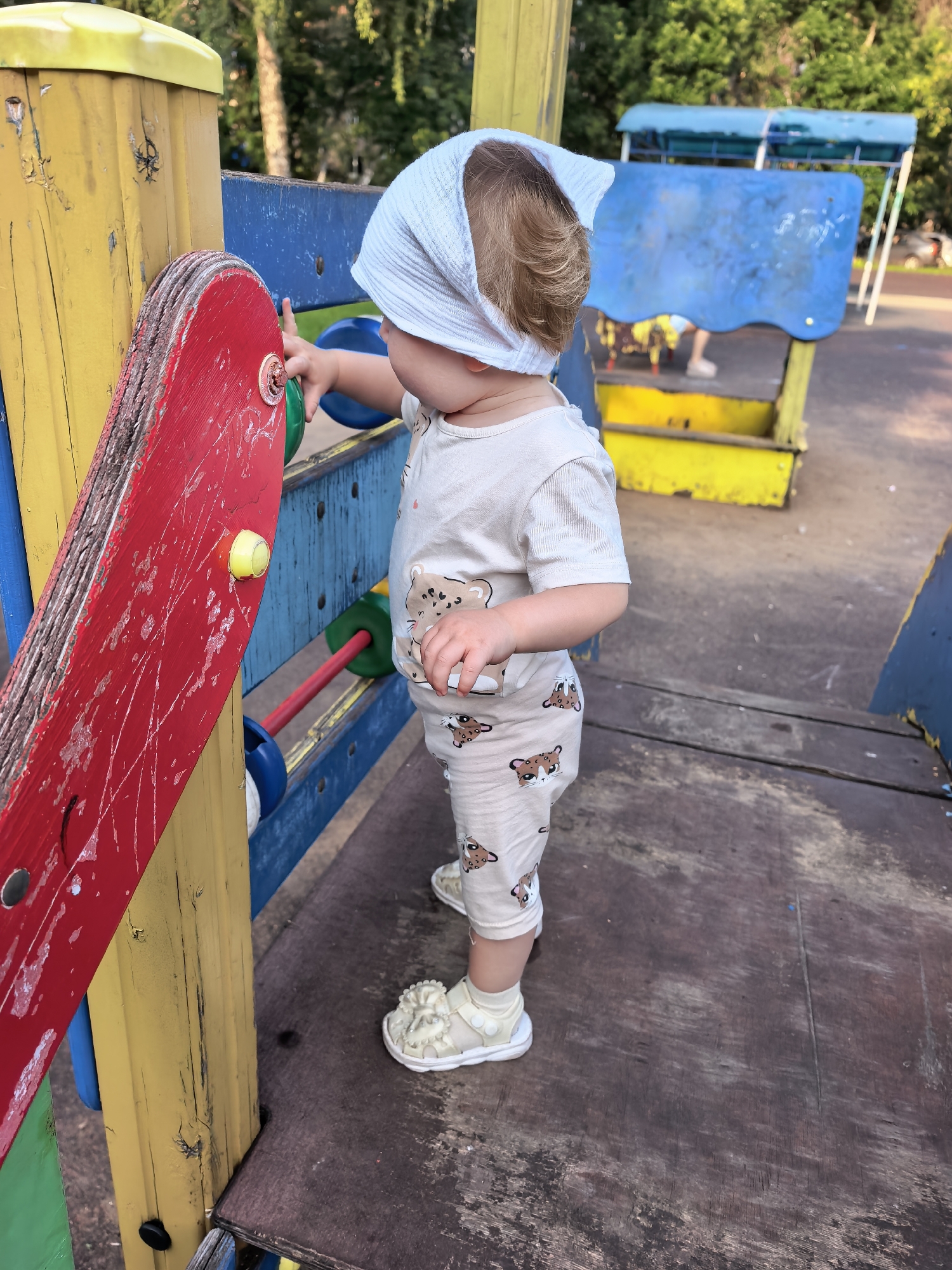A toddler, wearing a light-colored outfit with a pattern of bear faces and a white headscarf, stands on a wooden platform at a playground. The toddler is reaching out to touch a red, circular toy with green and yellow accents attached to a yellow and blue wooden structure. The playground equipment is brightly colored with red, yellow, and blue. In the background, trees and foliage are visible, suggesting an outdoor setting. The lighting indicates it is daytime, likely late afternoon given the warm light. The scene appears to be set in a park or recreational area in Sterlitamak, Russia. The toddler's posture suggests engagement and curiosity with the playground toy.