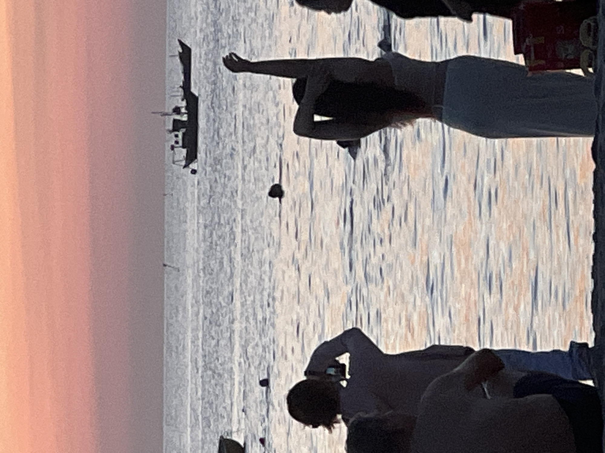 At sunset on a beach in Da Nang, Vietnam, several people are visible. In the foreground, a person is sitting with their back to the camera, wearing dark shorts. To their right, a person stands with their back to the camera, wearing a light-colored top and blue jeans, holding a camera to their face. Further to the right, a person stands facing away from the camera, wearing a white top and a long white skirt, with their arms raised above their head. Another person's head is visible to the far right. In the background, the ocean stretches to the horizon, with a pink and orange sky above. A small boat is visible in the distance on the water. Other people are present in the water, with heads visible above the surface. A red cooler is on the sand in the lower right corner.