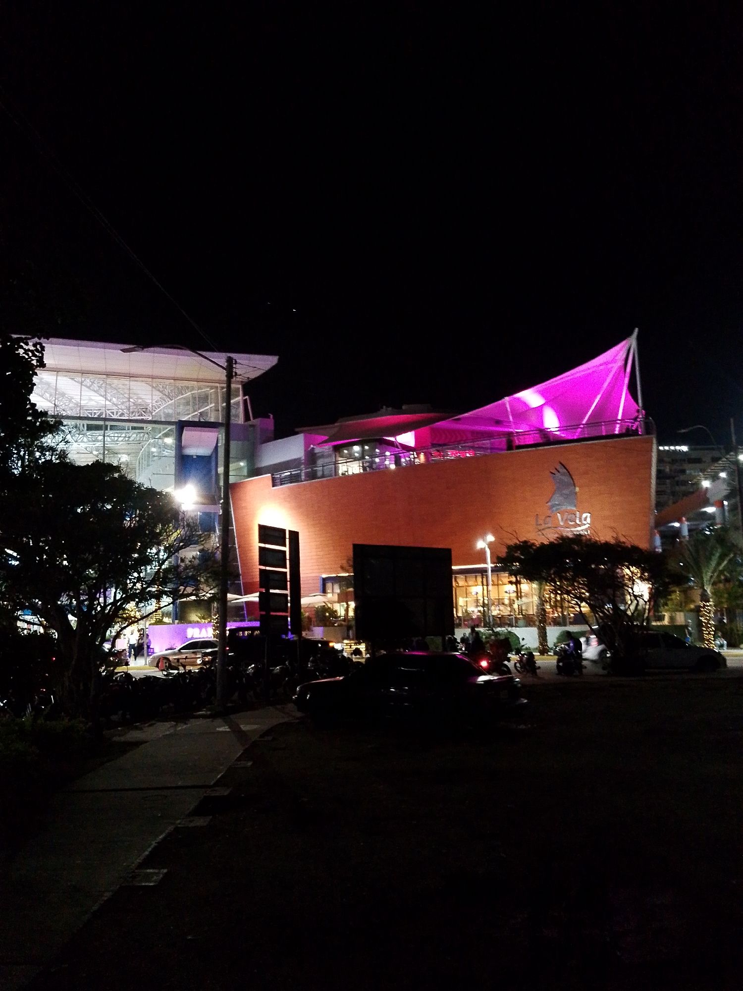This is a night-time, eye-level, medium shot of the "La Vela" shopping mall in Porlamar, Venezuela. The mall's facade is illuminated, with a prominent pink canopy structure creating a dramatic visual. The main building is brick-colored, with a large "La Vela" logo visible on its exterior. To the left, a modern glass and steel structure is also illuminated, housing the mall's interior. The foreground consists of a paved walkway and a dark grassy area. Several cars and motorcycles are parked and moving on the street in front of the mall, indicating activity. A few trees are present on the left side of the image, with their silhouettes dark against the illuminated buildings. The sky is dark, consistent with nighttime.