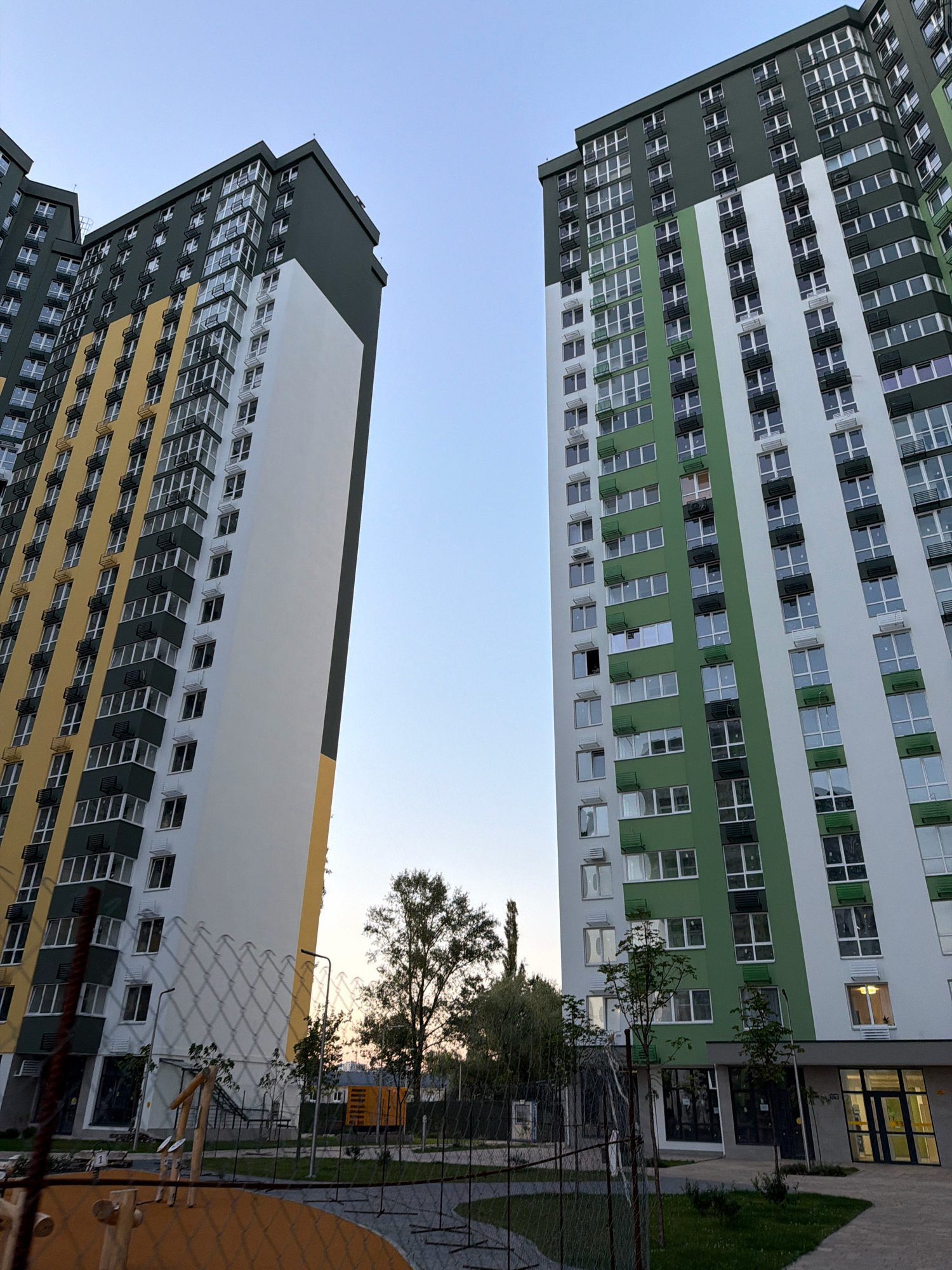 This is a medium-angle, eye-level shot of two modern, multi-story apartment buildings against a clear sky. The buildings are tall and feature a contemporary design with distinct color blocking. The building on the left is primarily white and yellow, with dark green accents and numerous windows with dark frames. The building on the right is white and green, also with dark green accents and a multitude of windows. Both buildings have visible balconies with dark railings and air conditioning units.
In the foreground, a chain-link fence runs horizontally across the bottom of the image. Beyond the fence, a small section of a playground is visible with orange and brown surfaces and wooden play structures. There are also streetlights and trees scattered between the fence and the buildings. In the background, between the two buildings, a yellow structure that resembles a construction trailer or modular unit can be seen. The lighting suggests it is either early morning or late evening, with soft light illuminating the scene. The image was taken in Kyiv, Ukraine.