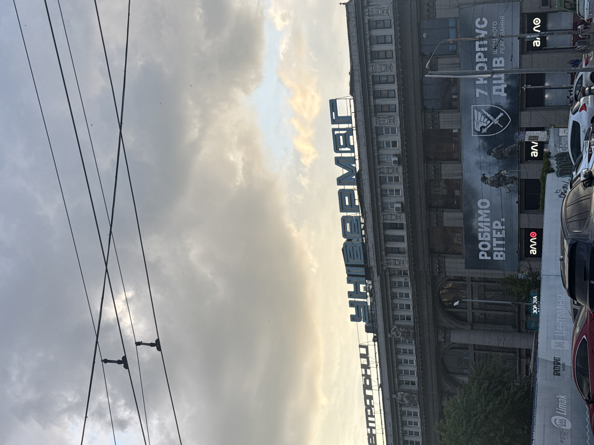 The image shows a building with the sign "УНІВЕРМАГ" (Univermag, meaning Department Store) in large blue letters across the top. The building appears to be in the city of Dnipro, Ukraine. Below the "УНІВЕРМАГ" sign, there is a banner with images of two soldiers and text in Ukrainian. On the lower part of the building, there are windows and decorative elements. In front of the building, there are several cars parked. The sky is cloudy, with some yellowish light suggesting either dawn or dusk. Overhead, there are electrical wires with insulators.