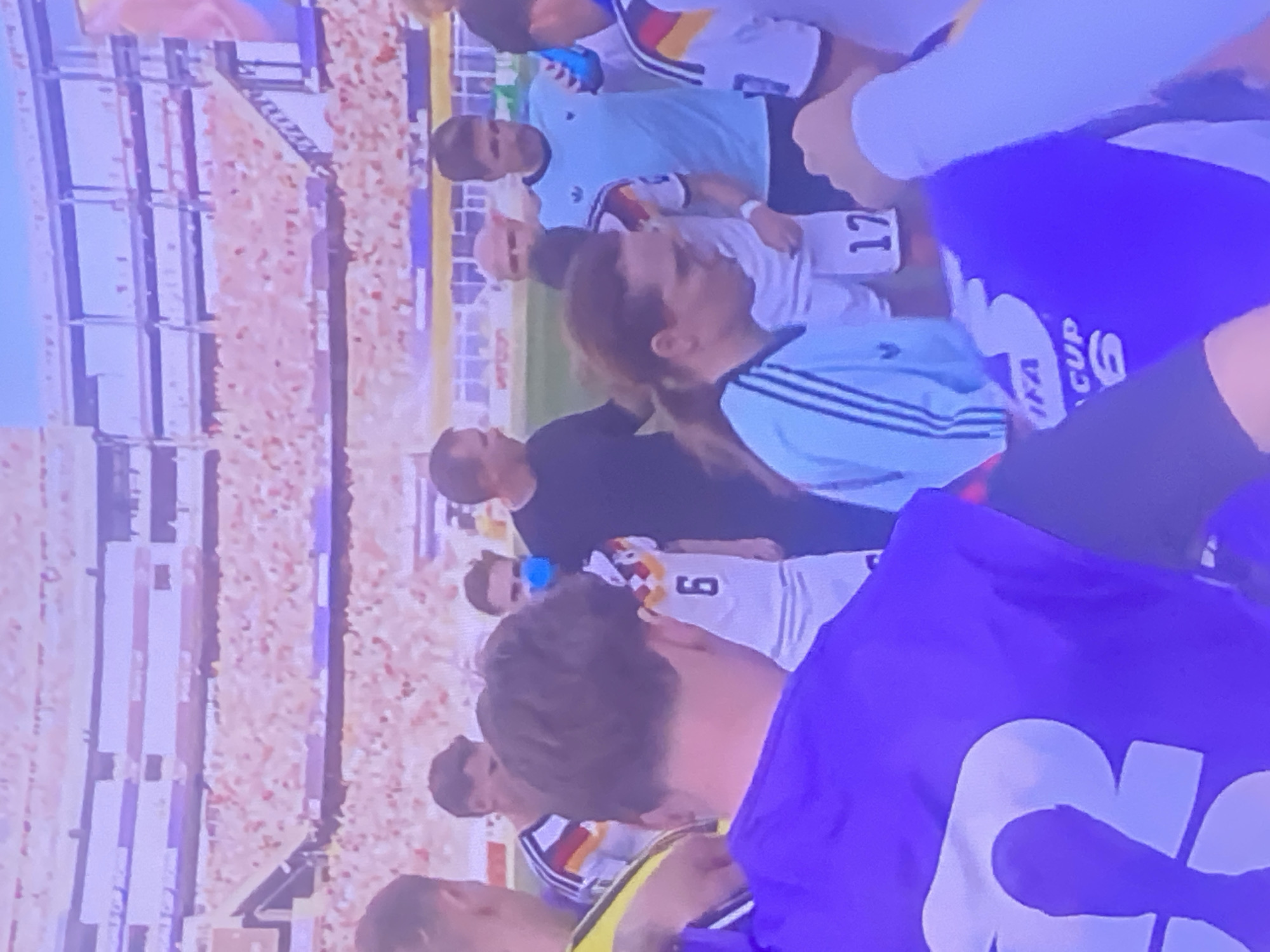The image shows a group of people in what appears to be a stadium setting with a blurred background of spectators. In the foreground, a person wearing a purple garment with a large white number "3" on it is partially visible. Another person is wearing a white jersey with the number "6" and a German flag motif on the sleeve. Several other individuals are visible, some in athletic wear, and one person is wearing a light blue shirt with stripes. A banner or flag with "FIFA CUP 6" is also visible. The overall impression is of a sports event, possibly a soccer match, with players and possibly staff members gathered. The city of Zastavna, Ukraine is not identifiable in this image.