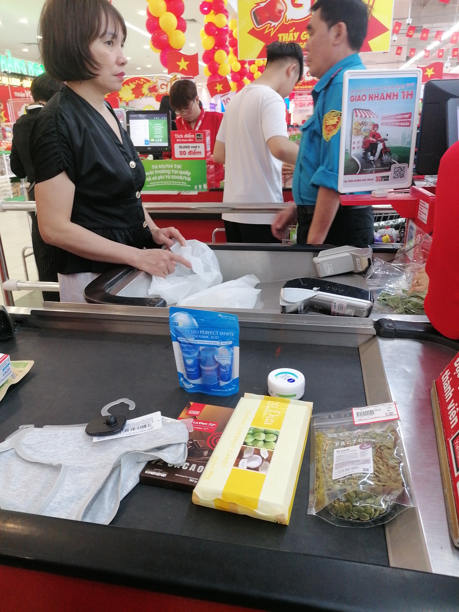 The image shows a checkout counter in a retail store, likely a supermarket, in Ha Long Ward, Vietnam, as evidenced by the Vietnamese text on signs and product packaging. A woman in a black blouse is standing by the counter, placing items into a white plastic bag. Other individuals are visible in the background, including a cashier in a red shirt and a person in a blue uniform. The items on the conveyor belt include a grey panty, a dark chocolate bar, a package of durian coconut candy, a bag of pumpkin seeds, a small blue bottle of facial treatment, and a Nivea cream. A sign promoting a "fast delivery in 1 hour" service is visible on the right. Overhead are red and yellow balloons, possibly indicating a promotional event or store opening.