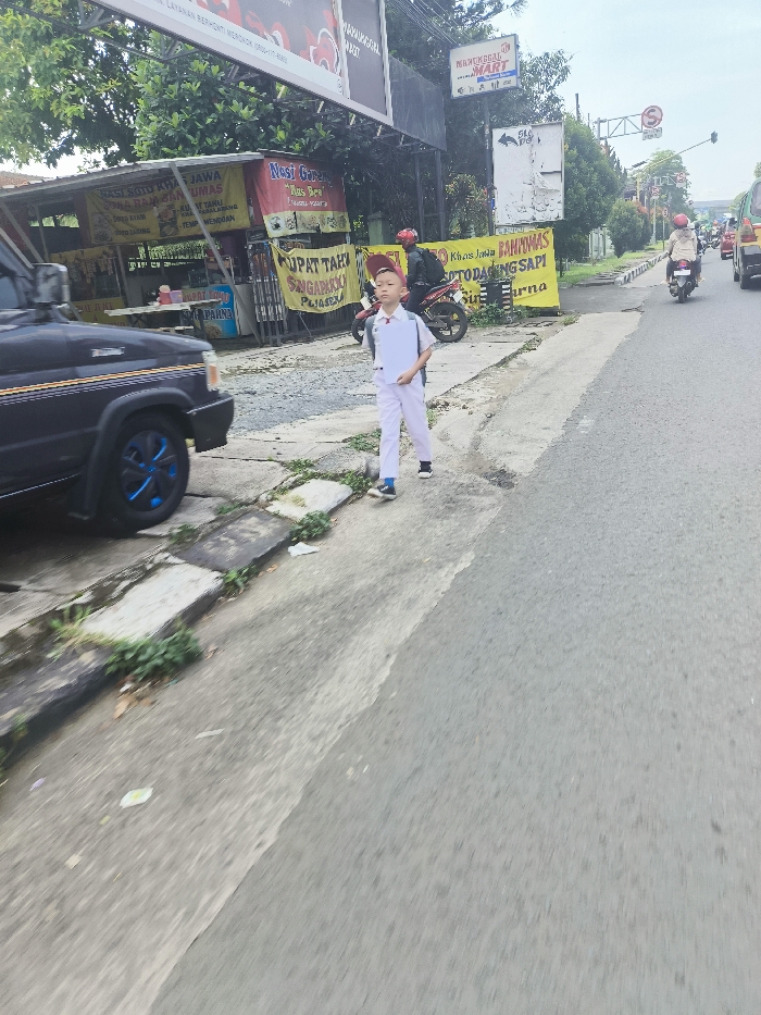 A young boy wearing a white uniform and a red cap walks along the side of a paved road in the town of Ngamprah, Indonesia, at coordinates -6.864, 107.512. He is walking on a narrow sidewalk next to a larger black SUV. The road is clear and the sun is out. The boy is carrying a white book. There is a man in a red shirt and a motorcycle parked behind the boy. The background shows several buildings, shops, and signs. There are also a few trees and some vegetation on the side of the road. A sign for "Bakwan Udang" is visible on the left. Another sign says "Khusus Jawa" is also visible. The background of the scene is a clear blue sky. There is a stop sign visible in the background. There are other cars and motorcycles visible in the distance.
