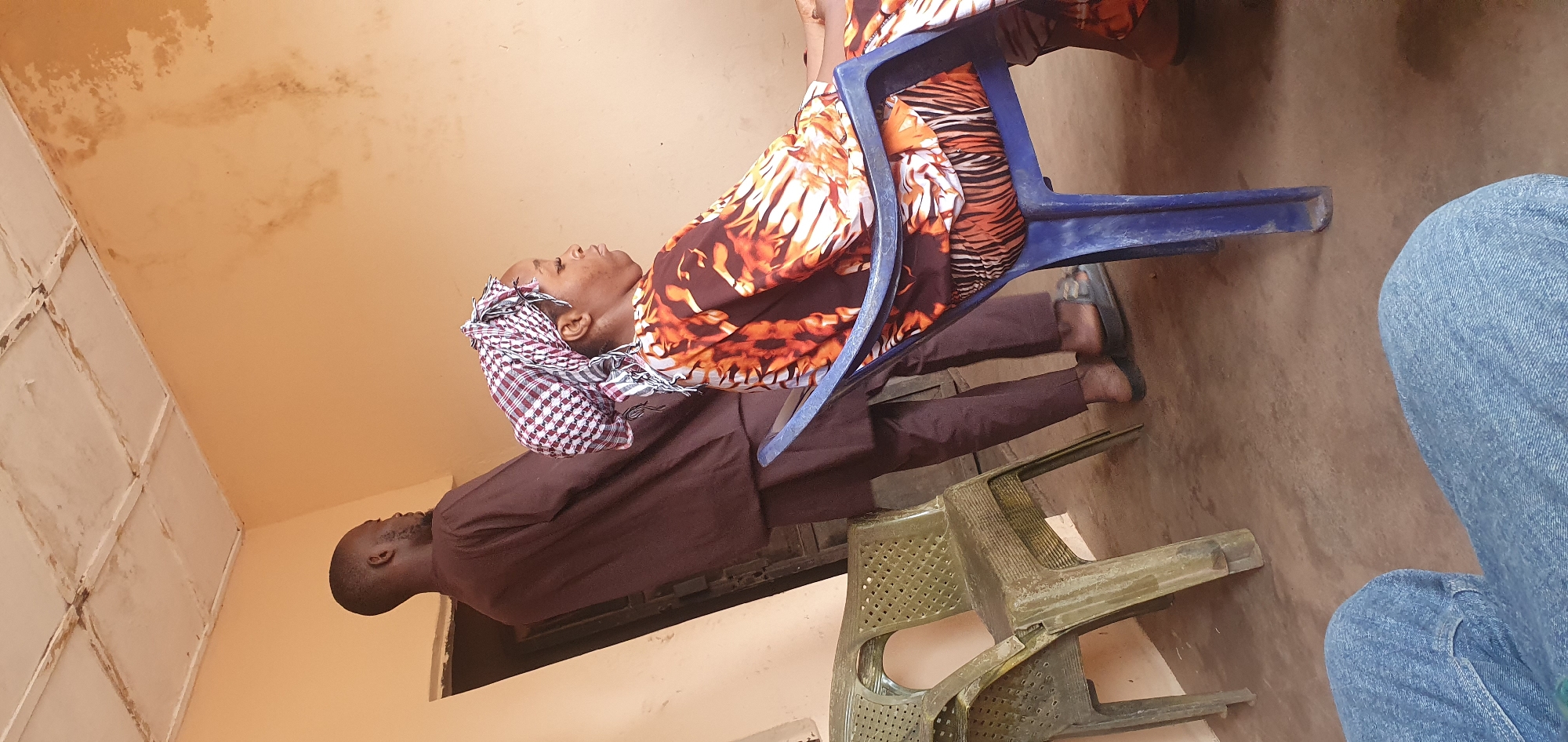 The image shows an indoor scene featuring three individuals. In the foreground, positioned centrally, a person is seated on a blue plastic chair. They are wearing a colorful, patterned long-sleeved top, possibly with an animal print design (orange, white, black, and brown), and dark brown trousers. A checkered head covering in red, white, and black fabric is wrapped around their head. Their bare feet are visible, wearing dark-colored sandals. This person is looking upwards and to the left.
Behind the seated individual, another person stands, facing away from the camera and slightly to the left. This person is dressed in a plain, long-sleeved dark brown garment, possibly a traditional tunic, and dark trousers. Only their profile and back are clearly visible.
In the bottom right corner, the legs of a third person, wearing blue jeans, are partially visible, suggesting another person is seated out of frame, possibly the photographer.
The setting appears to be a modest room with light beige-colored walls. One wall on the left side features a grid-like pattern, possibly molded plaster or paneling, showing some wear and discoloration. The ceiling, also light beige, exhibits significant dark brown stains and patches, indicating water damage. The floor is a reddish-brown, possibly concrete or dirt. A partially broken or inverted greenish-brown plastic chair is lying on the floor in front of the standing person. No specific activity or event is clearly depicted beyond the individuals being present in the room. There is no visible text. The overall scene suggests a casual indoor gathering in a simple, unadorned environment.