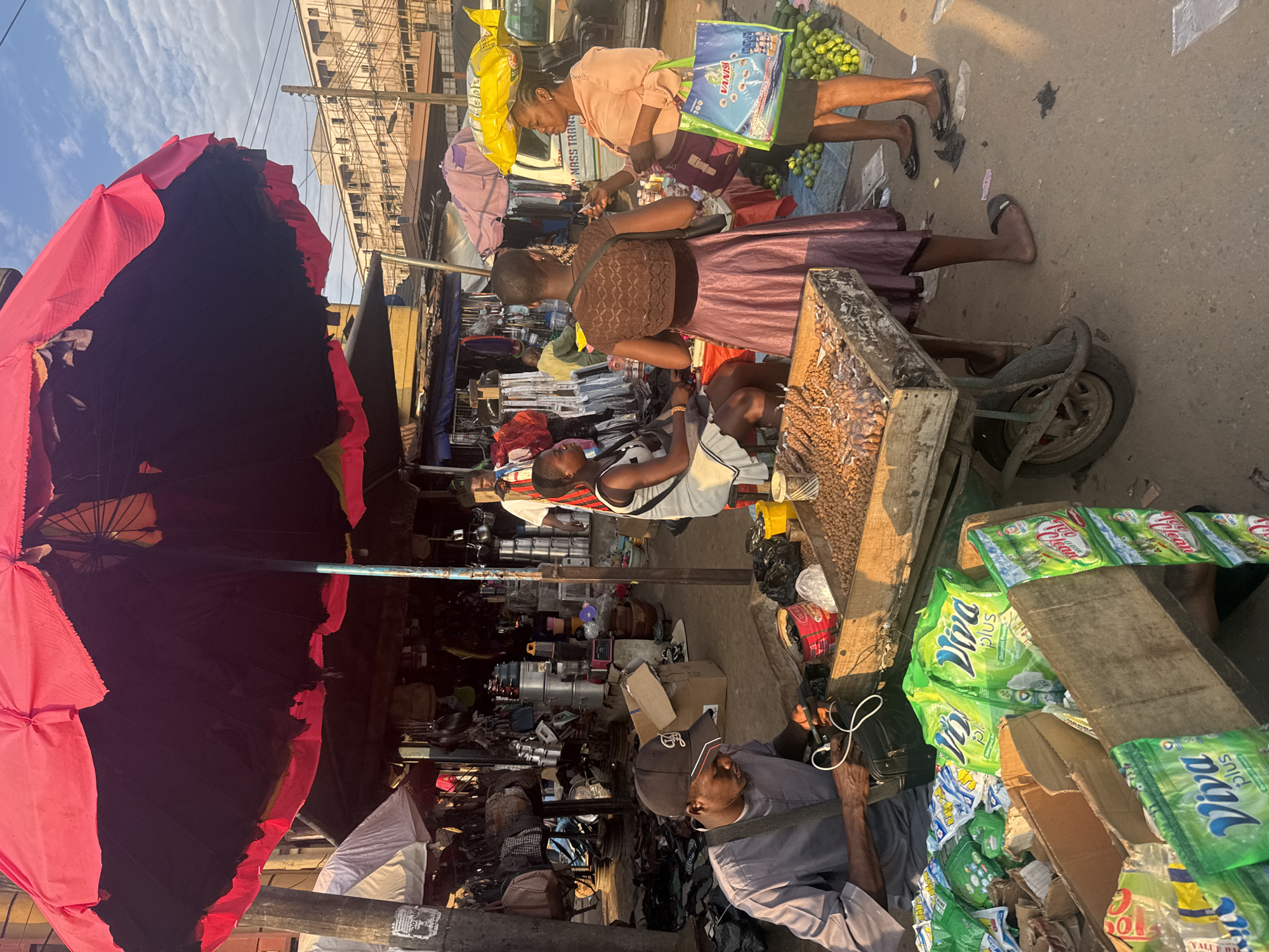 This is a street scene in Onitsha, Nigeria, likely within the bustling marketplace. A large, tattered red umbrella shades several vendors and customers. In the foreground, a man wearing a grey shirt and a black baseball cap is seated and appears to be charging his phone. He is surrounded by boxes of green detergent packets, some with the brand name "Viva Plus" visible.
To his right, a wooden cart displays a large quantity of dried goods, possibly nuts or seeds. Behind the cart, three women are engaged in transactions. One woman, wearing a pink dress, is standing and holding a blue and green shopping bag. Another woman, in a brown top and purple skirt, faces away from the camera. A third woman, seated and wearing a white dress, is also visible.
In the background, stalls are set up with various goods, including pots, pans, and other household items. A white truck with the text "MASS TRANS" is parked behind the stalls. The ground is paved with concrete and appears to be a well-trafficked area. The lighting suggests it is either early morning or late afternoon, with the sun casting long shadows. The overall atmosphere is that of a busy, active marketplace.