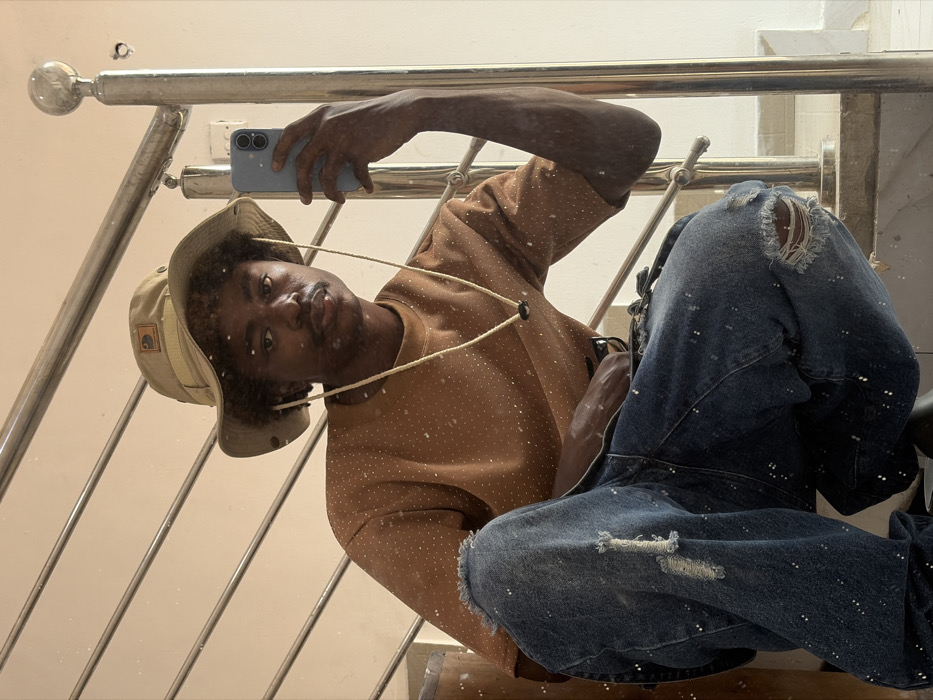 The image captures a young Black male crouching indoors in Igboefon, Nigeria, reflected in what appears to be a mirror covered with fine white specks, possibly dust or paint. He is actively taking a selfie using a light blue smartphone held in his raised left hand. The individual wears a tan boonie hat with a chin strap, a muted orange crew-neck short-sleeved t-shirt, and distressed blue denim jeans with large rips at both knees, all of which are visibly splattered with small white paint marks. He has a short afro hairstyle and a faint mustache, maintaining a neutral expression directed towards the camera. The background, visible through the reflection, consists of a light-colored wall and a multi-barred metallic staircase railing.
