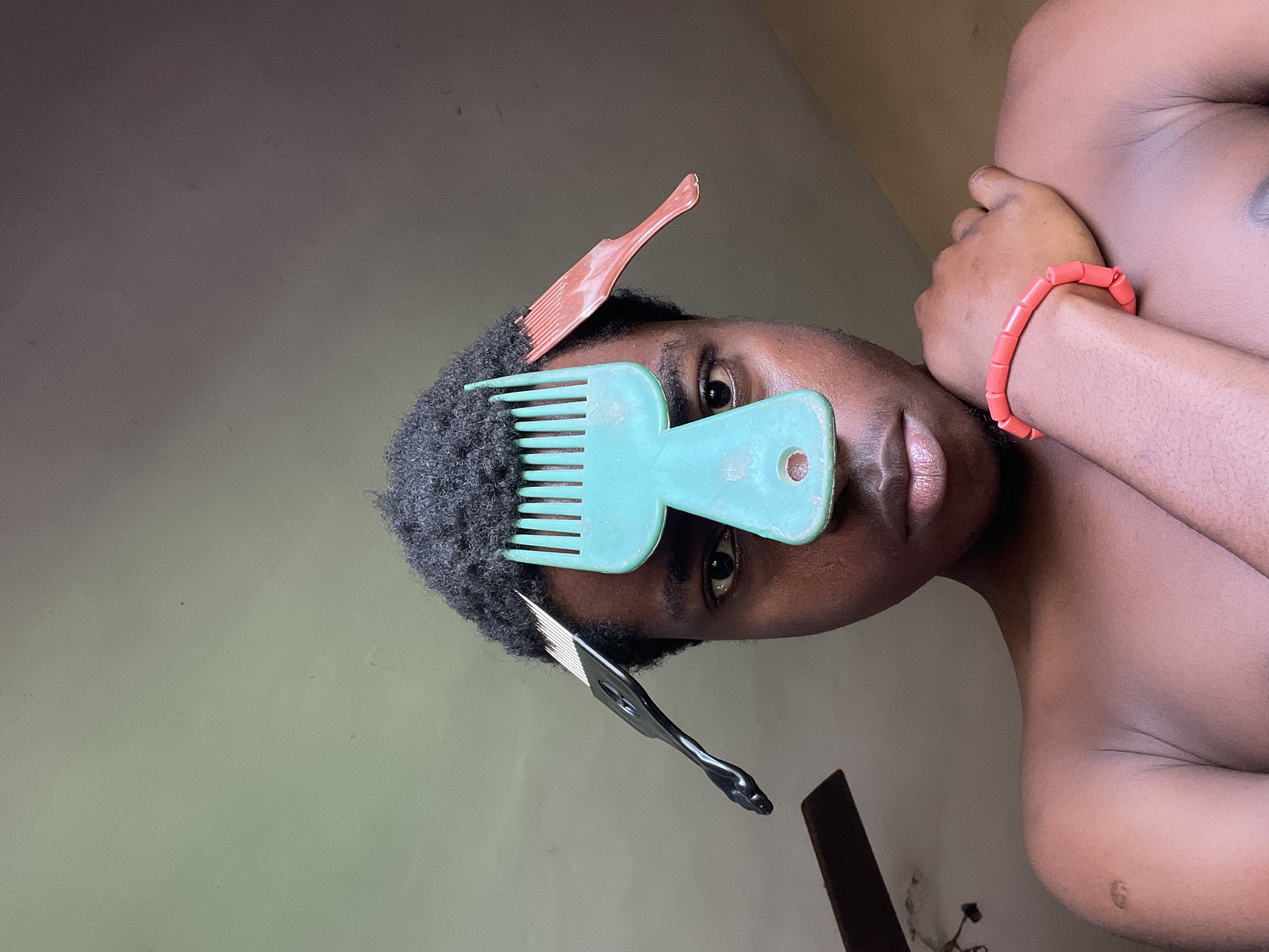 A close-up, rotated, and slightly upward-angled shot shows a shirtless person with dark skin and short, curly black hair in Nkpor, Nigeria. Three combs are inserted into the person's hair: an orange comb near the top, a blue comb covering one eye, and a black comb on the lower left side. The person has dark eyes and a serious expression. The person's right arm is bent, with a red beaded bracelet visible on the wrist. The background is a mottled grayish-green wall. A portion of a ceiling fan is visible at the bottom of the image.