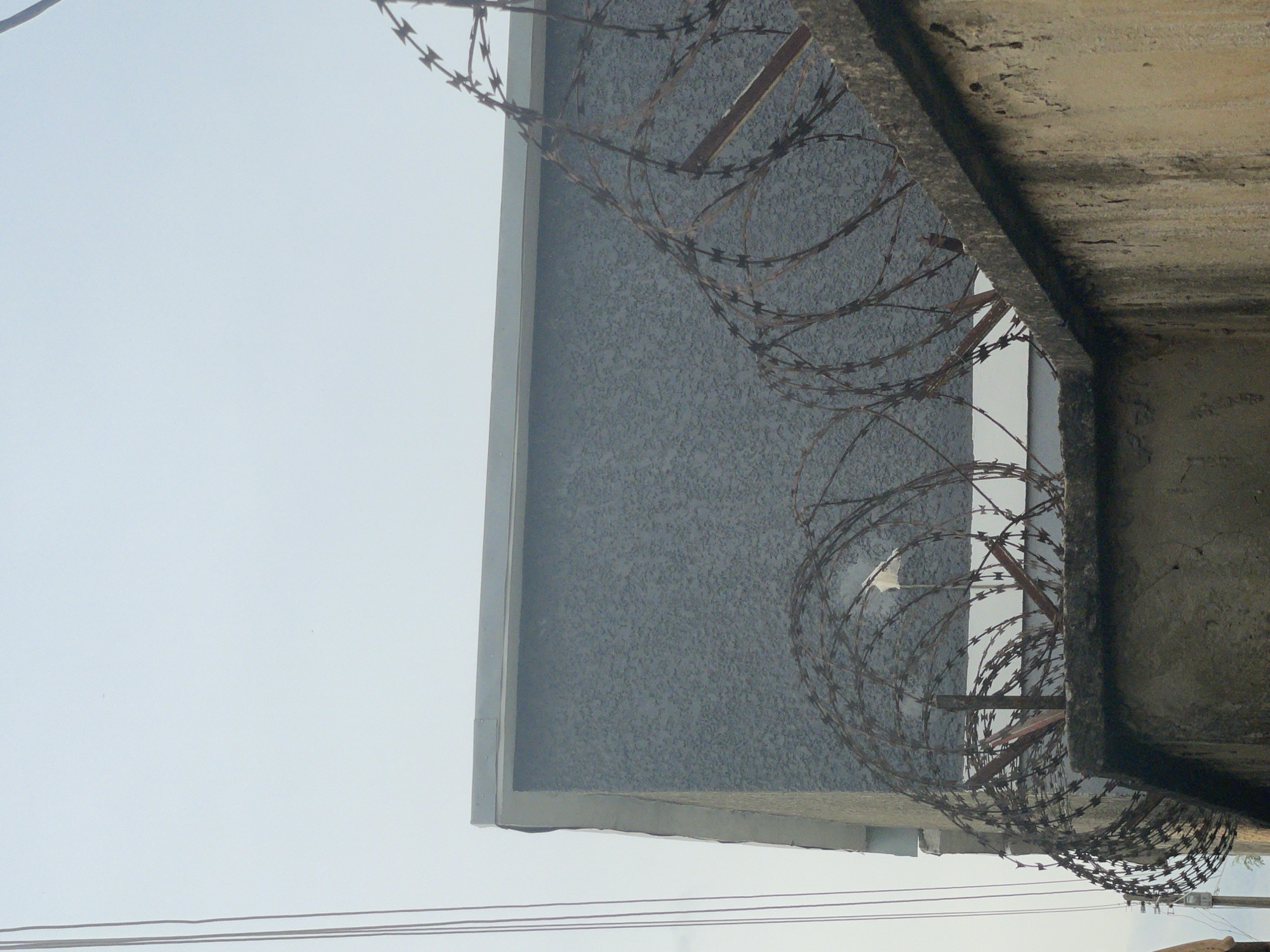 A concrete wall topped with barbed wire fences a building with a gray, textured exterior. The sky above is overcast. Several thin wires, possibly electrical or telephone lines, run vertically along the left side of the image. The scene appears to be an outdoor environment. The location is in the city of Narayi, Nigeria.