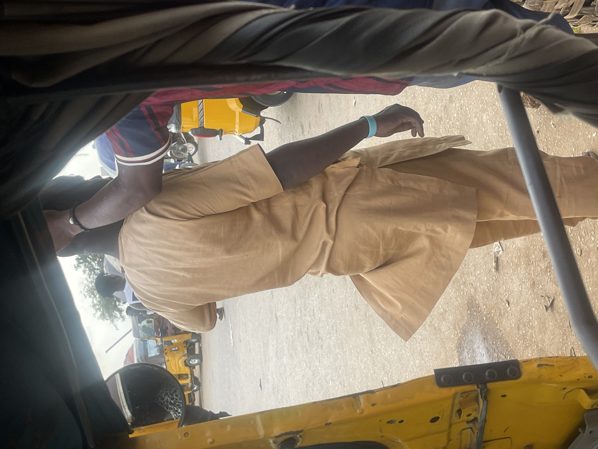 The image, captured within the bustling environment of Galadimawa Village, Nigeria, depicts a street scene dominated by a yellow auto-rickshaw, locally known as a "Keke." The foreground is occupied by the interior of another Keke, framing the view. A man, seen from the back and side, is dressed in a tan, loose-fitting traditional outfit, with a bracelet on his left wrist. He appears to be interacting with someone to his right, whose arm, adorned with a striped sleeve, is visible. In the background, other Kekes and individuals are discernible, suggesting a typical day in this Nigerian locale. The ground is unpaved, indicative of a rural or semi-urban setting.
