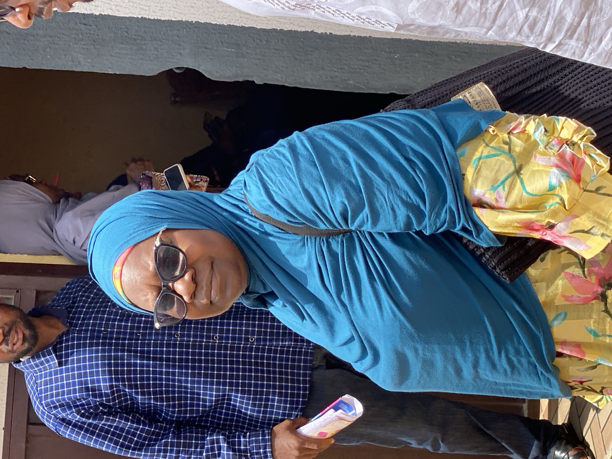 A medium shot of a woman wearing a blue hijab and sunglasses, standing next to a man in a blue checkered shirt, holding a rolled-up stack of papers. A yellow floral print bag is seen behind her. The location is identified as Sabon Tasha, Nigeria.