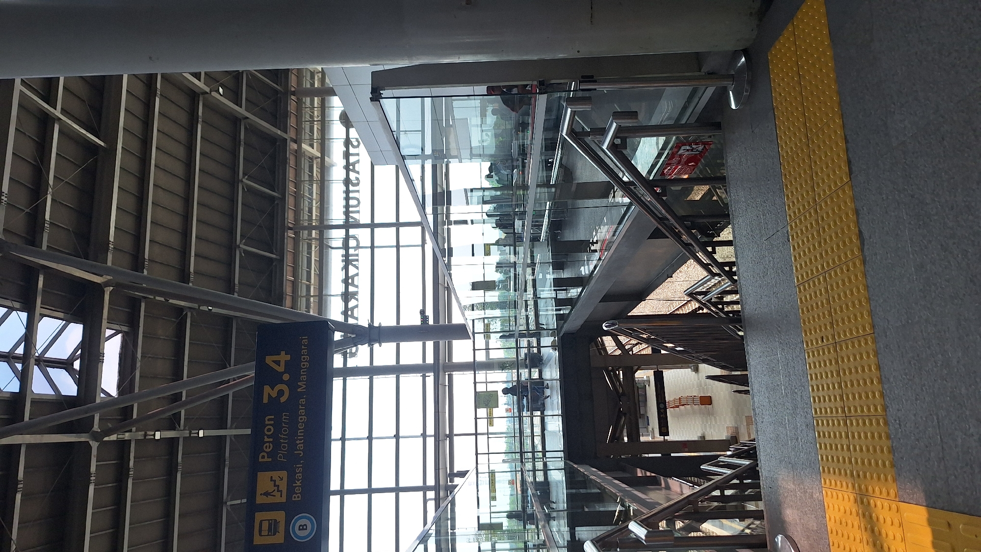 The image depicts the interior of Cikarang Station in Cikarang, Indonesia. The view is angled upwards. The building is constructed with a visible steel frame and a paneled roof. A blue sign indicates "Peron (Platform) 3,4" with destinations "Bekasi, Jatinegara, Manggarai," with icons for train access and bus access. A glass-enclosed overhead walkway is visible, with silhouettes of people inside. The floor has a yellow tactile paving strip for visually impaired individuals. Stairs with steel railings are present, leading to a lower level.