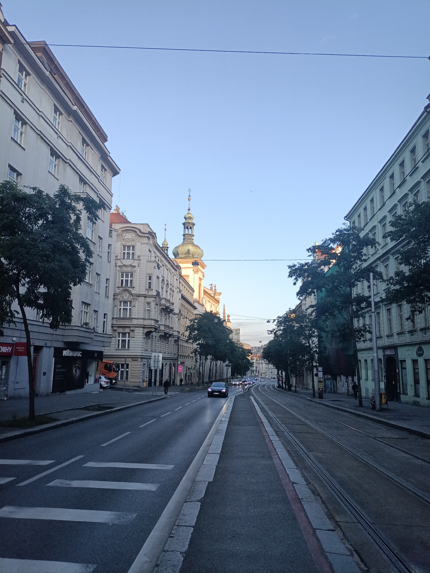 The image depicts a street scene in Prague, Czechia, likely in the Old Town or a similar historic district. The perspective is from the street level, looking down a wide avenue lined with multi-story buildings. The architecture suggests a European city with a mix of styles, including ornate facades on some buildings and simpler, more modern designs on others.
On the left side of the street, a light-colored, multi-story building with numerous windows dominates the foreground. A mature tree with dark foliage stands on the sidewalk in front of it, partially obscuring the lower floors. Various signs are visible on the ground floor, including one for "PRISM OPTIC" which features a large eye graphic. An orange truck is parked behind a pedestrian crossing.
The right side of the street features a similar row of buildings, with the one in the foreground painted a pale green and appearing to have a more repetitive window pattern. Trees are also present along this side, providing a canopy over the sidewalk in places.
The street itself is paved and shows signs of wear. Prominent tram tracks run down the center of the road, indicating public transportation is a key feature of the area. A section of a pedestrian crossing with white stripes is visible in the foreground on the left side of the road.
Several vehicles are present: a dark-colored car is driving away from the camera in the middle of the road, and another dark-colored car is visible further down the street. A few pedestrians can be seen on the sidewalks. The sky is a clear, pale blue, suggesting it is either early morning or late afternoon, with the sun casting long shadows. The overall impression is of a city awakening or winding down, with a blend of historical architecture and functional urban infrastructure.