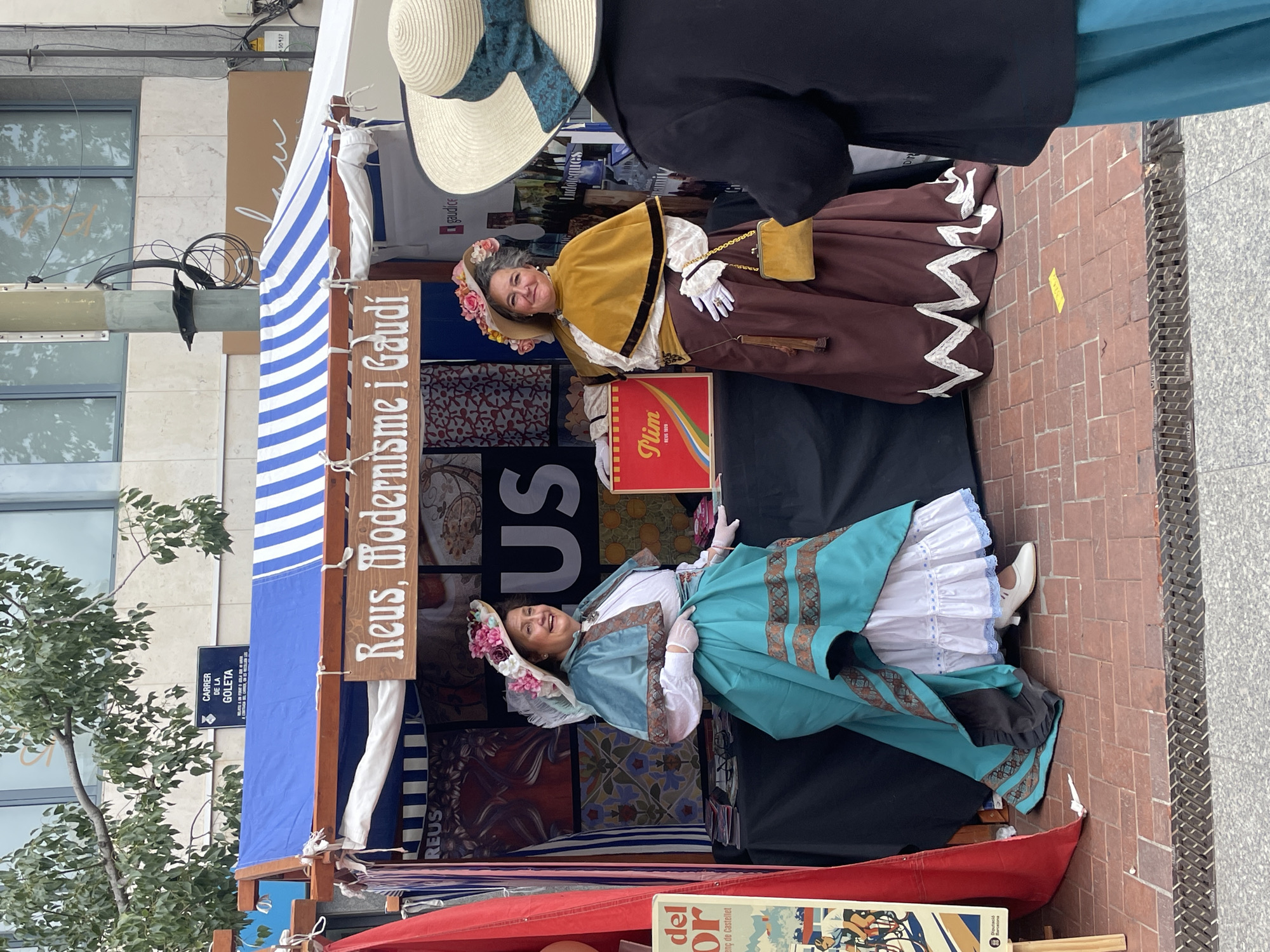 The image shows two women dressed in vintage or historical costumes standing behind a stall at a market or fair. A sign reads "Reus, Modernisme i Gaudí," suggesting a theme of modernism and the architect Antoni Gaudí. The stall is decorated with blue and white striped fabric. Another sign on a nearby building reads "CARRER DE LA GOLETA." A red box labeled "Plim" is visible on the stall, as is a poster with the words "del or." Based on the street name and the reference to Reus, this likely takes place in or has connections to Reus, Spain.