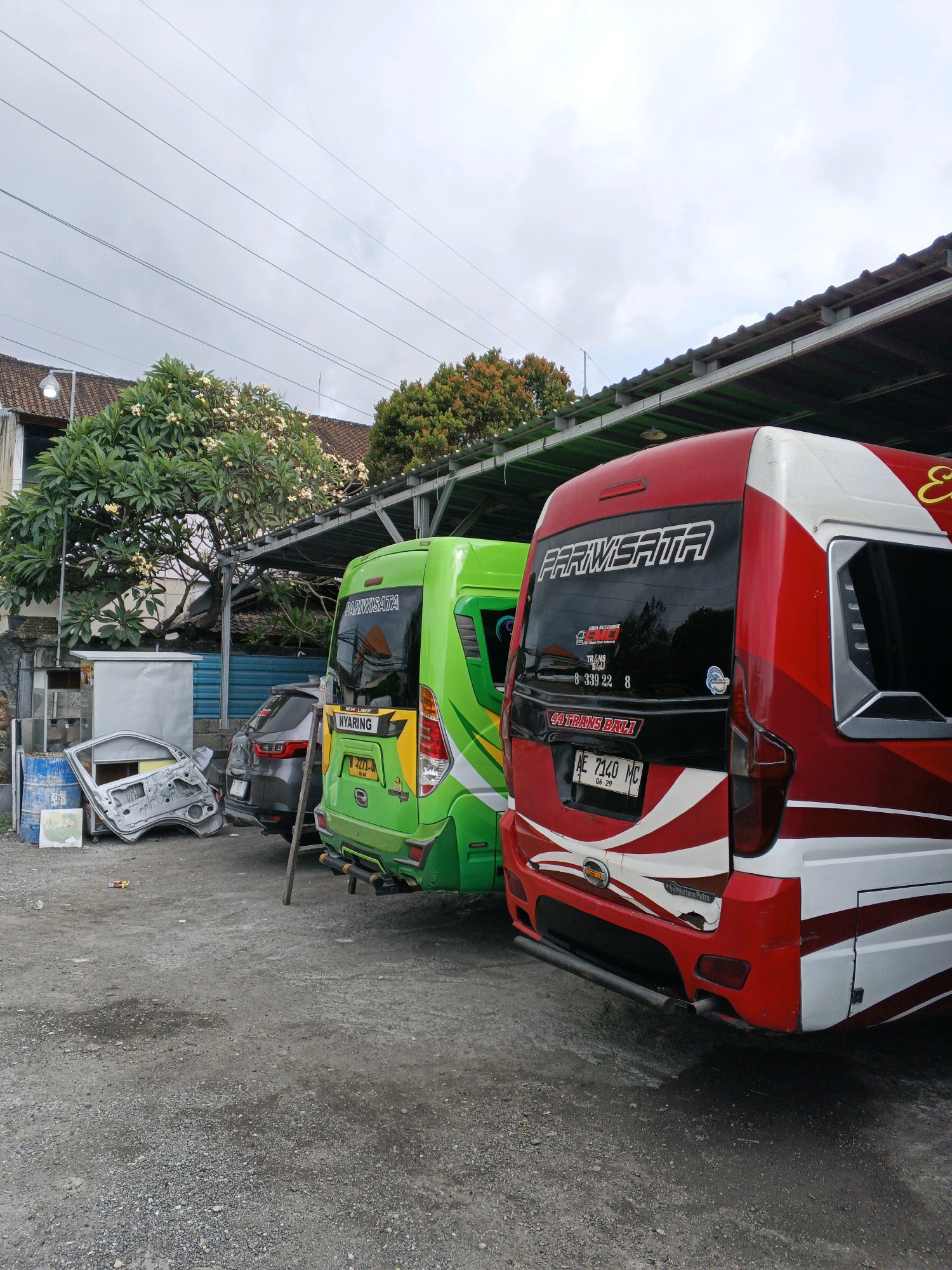 The image depicts a street scene in Denpasar, Indonesia, likely an area with automotive repair or parking facilities. Two buses are prominently parked side-by-side. The bus on the right is red and white with "PARIWISATA" and "44 TRANS BALI" written on its rear. It has the license plate AE 7140 MC. The bus on the left is bright green, also displaying "PARIWISATA" and "NYARING" on its rear, with the license plate E 3275 J. In front of the buses, to the left, is a detached car door leaning against a blue barrel and a metal storage unit. A dark gray car is parked between the buses and the detached car door. The environment appears to be an outdoor lot with a gravel and asphalt surface, shaded by a corrugated metal roof structure supported by metal poles. Trees with white flowers are visible in the background, along with a building with a tiled roof. The sky is overcast. There are no people visible in the frame. The scene suggests a place where vehicles are serviced, stored, or awaiting repair.