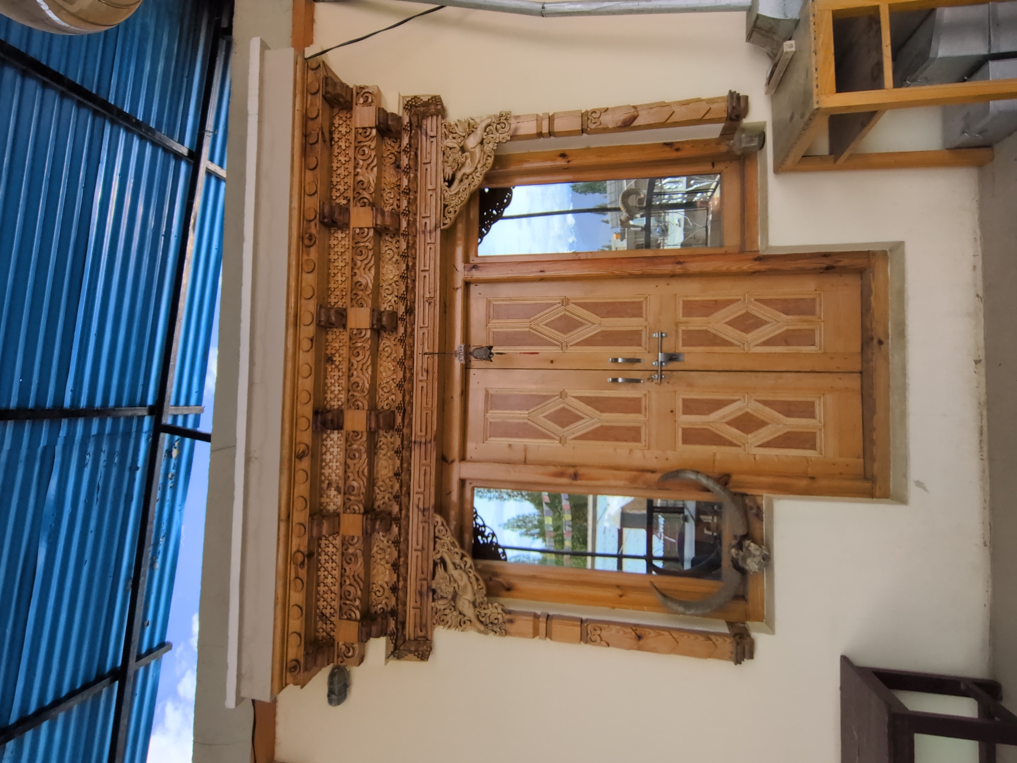 The image shows a wooden door with intricate carvings above it and on the side frames, flanked by two windows. The door has two panels with a diamond pattern and is secured with a latch. The left window displays a pair of animal horns and a skull. The right window reflects mountains and flags. Above the doorway is a decorative wooden structure with detailed carvings. A blue corrugated metal roof with supporting metal beams is visible overhead, along with a white ledge. In front of the door, on the left, is a dark wooden bench, and on the right, a light-colored wooden table with metal boxes underneath. The scene appears to be outdoors, possibly in the city of Leh, India, given the architectural style and the reflection of mountainous terrain. The walls are painted a light beige color.