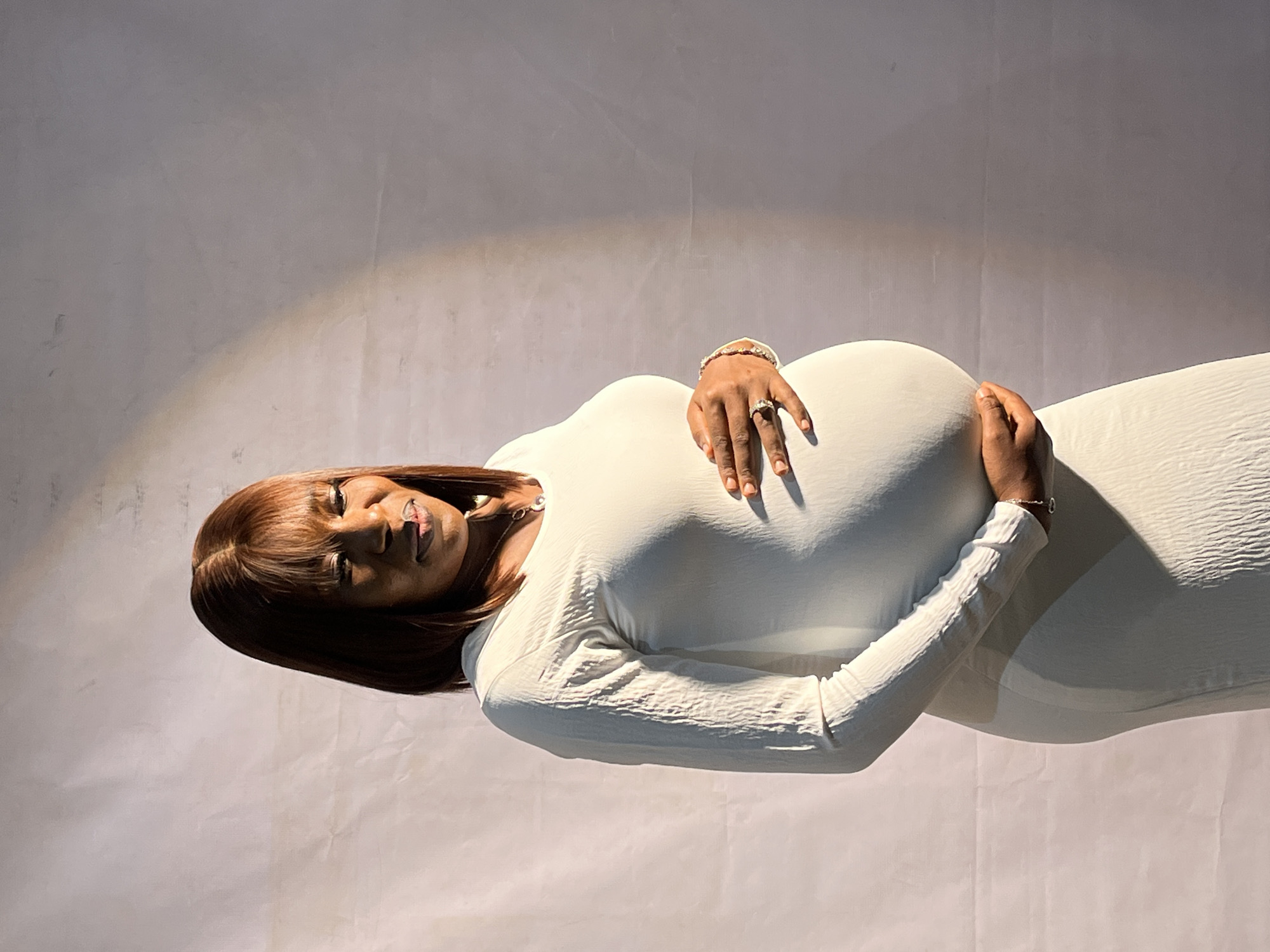 A medium shot captures a pregnant woman with fair skin, dark hair styled with bangs, and dark eyes. She is wearing a long-sleeved, light-colored dress with a textured pattern and a necklace. The woman is facing right, with her eyes closed and a soft smile on her face, her hands resting on her prominent belly. She is adorned with rings on her fingers and a bracelet on her wrist. The background consists of a light-colored, textured wall, with a stark shadow cast from the right side, creating a spotlight effect on the woman. The lighting suggests it is daytime. The scene is not specifically identifiable to Jos, Nigeria, based on visual cues alone.
