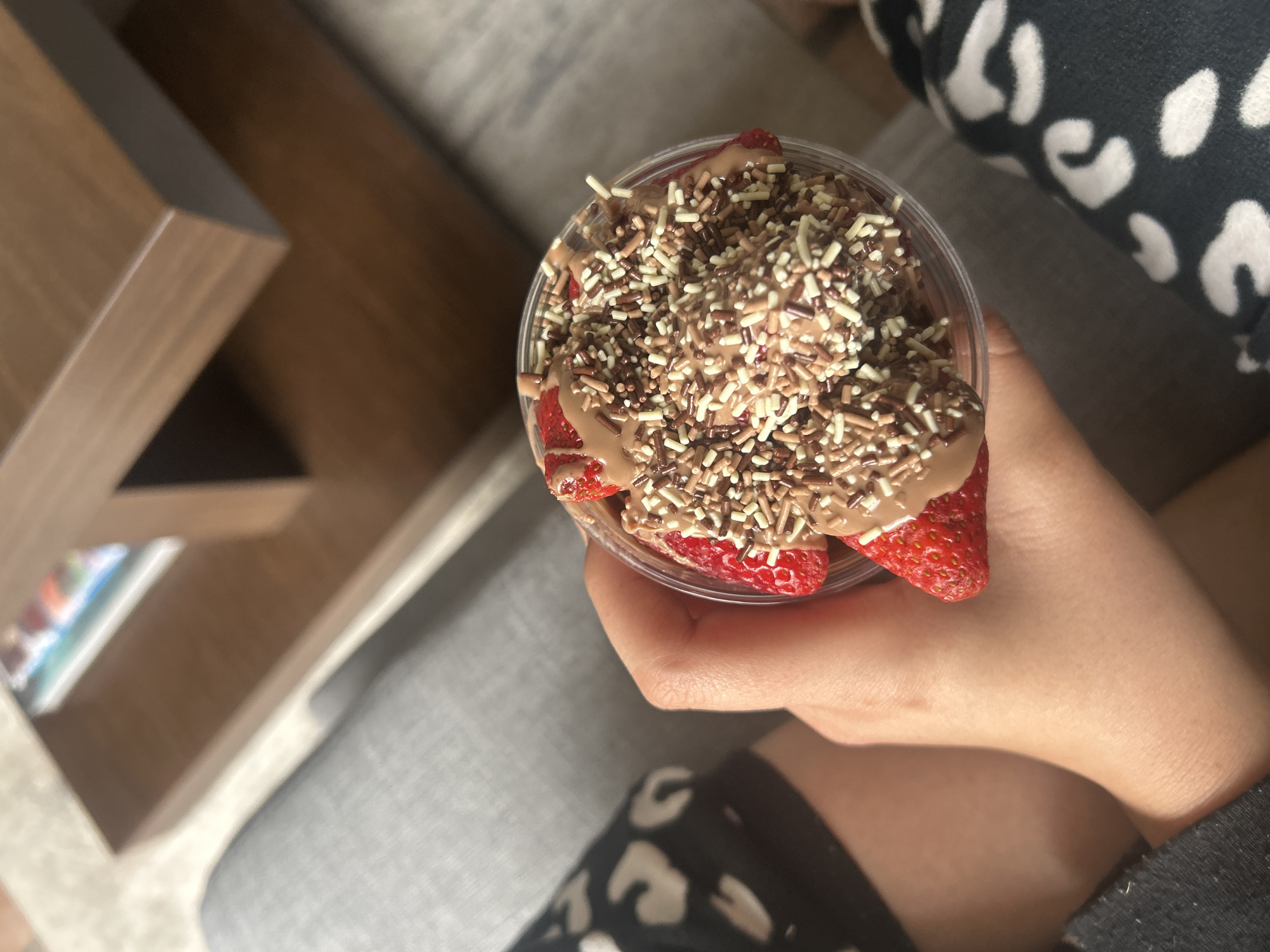 A high-angle shot reveals a person holding a clear plastic cup filled with strawberries, chocolate sauce, and sprinkles. The person wears patterned clothing. In the background, a section of a brown wooden shelf and a gray upholstered surface are visible. The location is Unknown, Unknown.