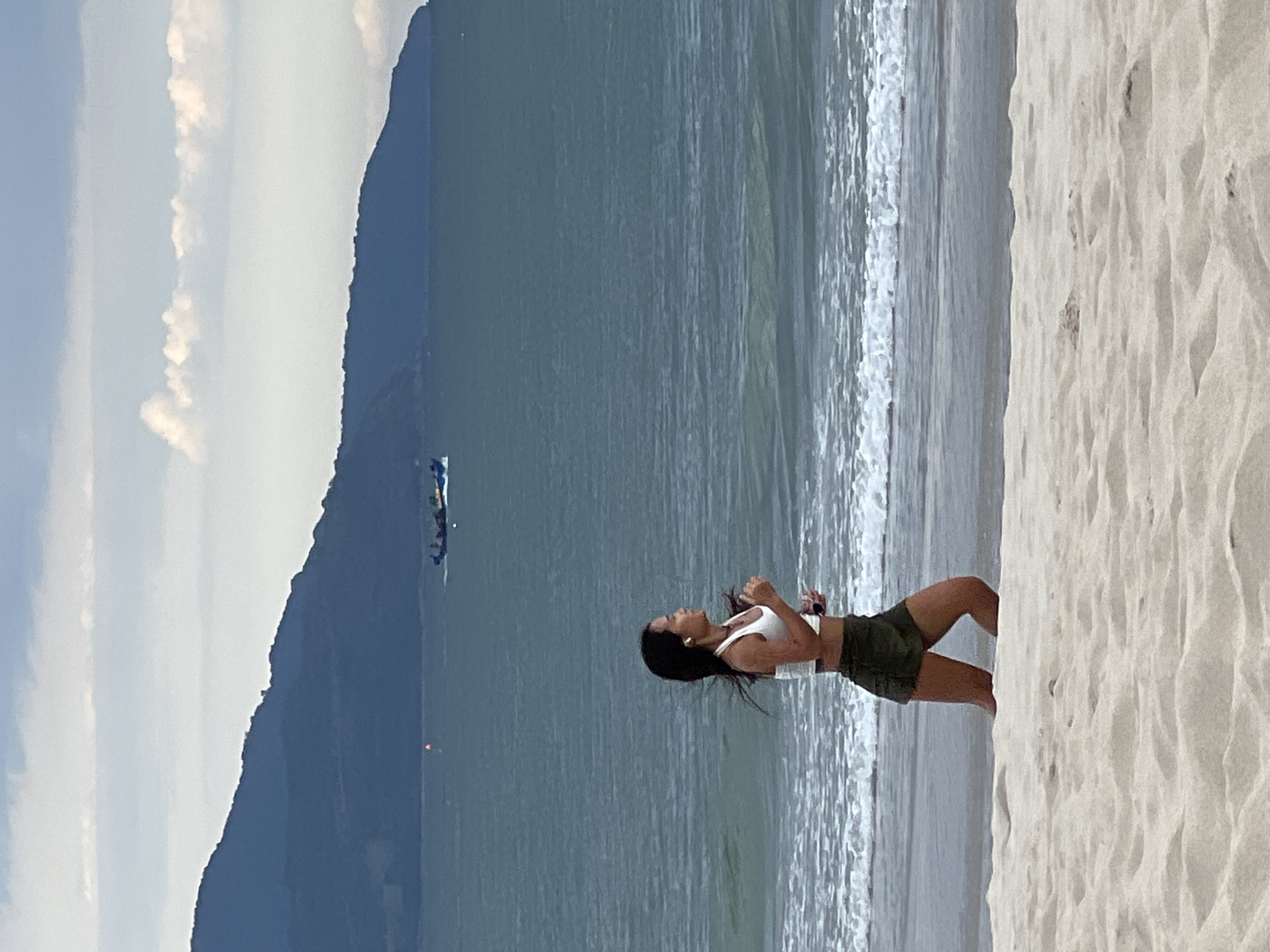 The image shows a woman standing on a sandy beach, facing the ocean. She is wearing a white crop top and olive green shorts. Her hair is dark and appears to be blowing in the wind. She is looking up and to her left, with her right arm raised. There are waves gently breaking on the shore in front of her. In the distance, across the water, a large, dark mountain looms, with clouds scattered across the sky above it. There is also a small boat visible on the water in front of the mountain. The sand in the foreground is light-colored and appears to have footprints on it. The lighting suggests it is daytime, likely late afternoon or early morning due to the soft light and the colors of the sky. The scene evokes a sense of peace and contemplation.