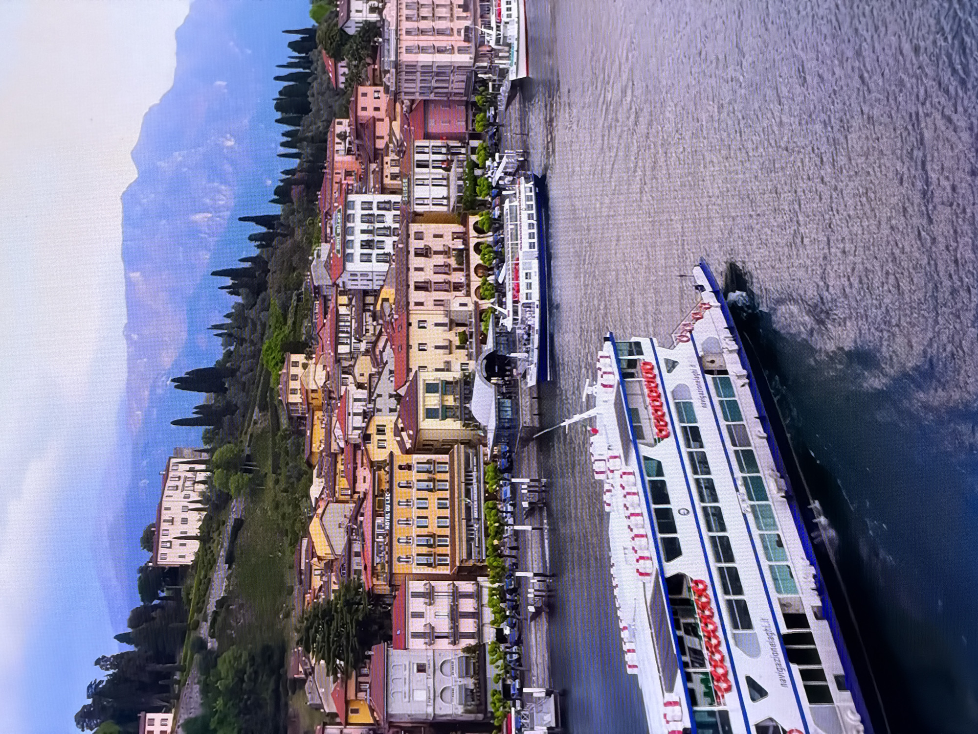 The image depicts a scenic waterfront with a town nestled on a hillside overlooking a body of water. The architecture of the town features buildings with terracotta roofs and pastel-colored facades, suggesting a European setting. Several boats are visible on the water, including a large white ferry in the foreground with blue accents and several life preservers visible along its side. Further back, other vessels, including a smaller white ferry, are docked or moving. The hillside behind the town is covered with lush greenery and trees, with a prominent building situated higher up. In the background, a majestic mountain range rises, shrouded in a soft, hazy light. The sky is a pale blue. The scene captures a tranquil, picturesque day at a lakeside or seaside town. This description does not align with the city of Chirchik, Uzbekistan, as the architectural style and natural landscape are not characteristic of that region.