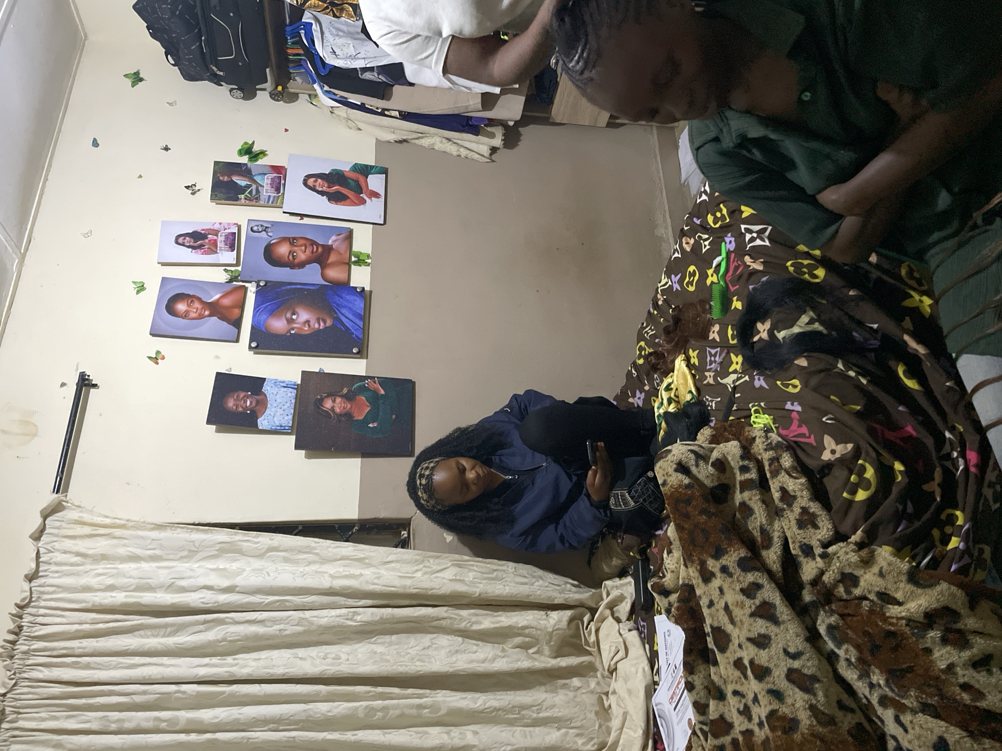 In a room in Durumi, Nigeria, a young woman with dark, curly hair sits on a bed covered with a leopard-print blanket and a brown blanket with a Louis Vuitton pattern. She is wearing a blue jacket and black pants and is looking down at her phone. On the wall behind her are several framed portraits of women. To her right, another young woman with braided hair, wearing a green shirt, is also sitting on the bed, her arms crossed. In the background, clothes hang on a rack, and luggage is stacked on a shelf. The room appears to be a bedroom.