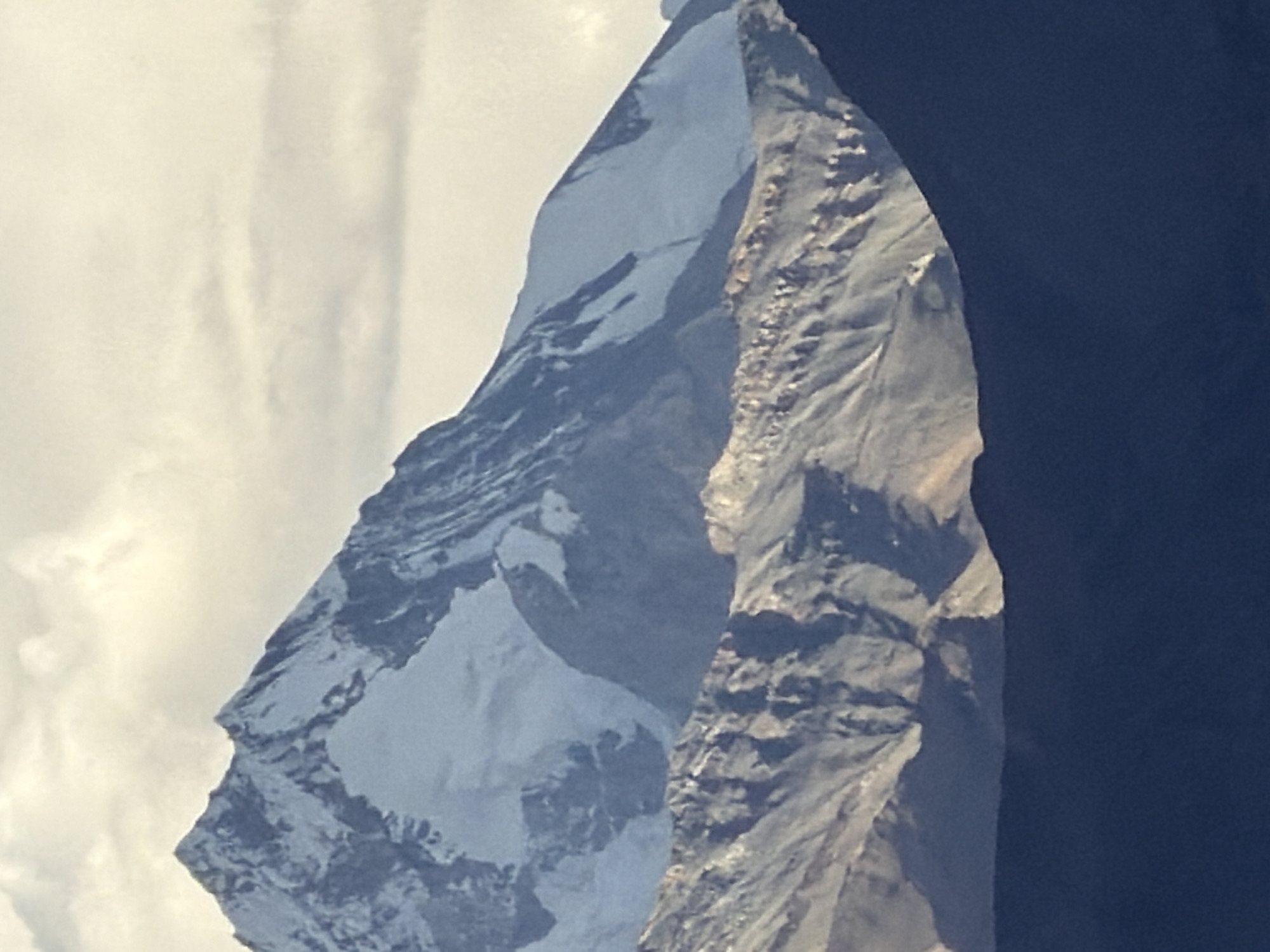 The image shows a majestic mountain range under a cloudy sky. The foreground features a dark, silhouetted mountain range. Behind this, a textured, rocky mountain slope is visible with shadows and highlights suggesting rugged terrain. The furthest and highest mountain peaks are snow-capped and appear imposing against the sky. The overall impression is of a grand, natural landscape. The location is identified as Unknown, India.