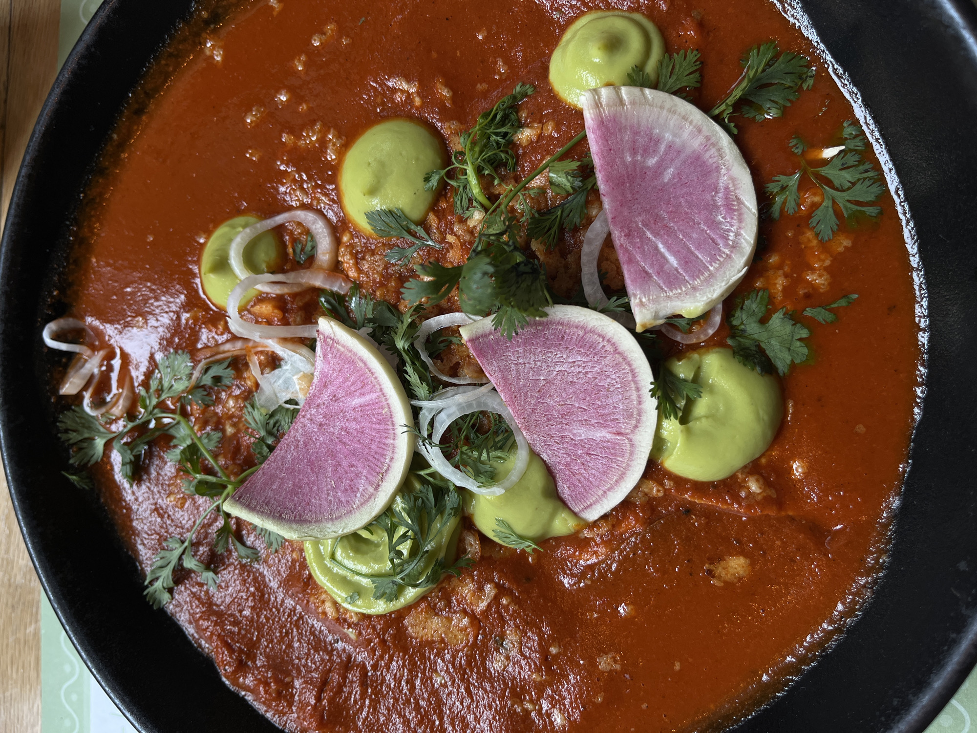 A close-up, overhead shot displays a plate of Mexican cuisine in San Bernardino Tlaxcalancingo, Mexico. The dish features a vibrant red sauce covering the base, which appears to contain enchiladas or a similar type of filled tortilla. Toppings include dollops of light green avocado cream, slices of pink-purple watermelon radish, thin slivers of white onion, and sprigs of fresh cilantro. The dish is served in a deep, black, possibly cast-iron pan.