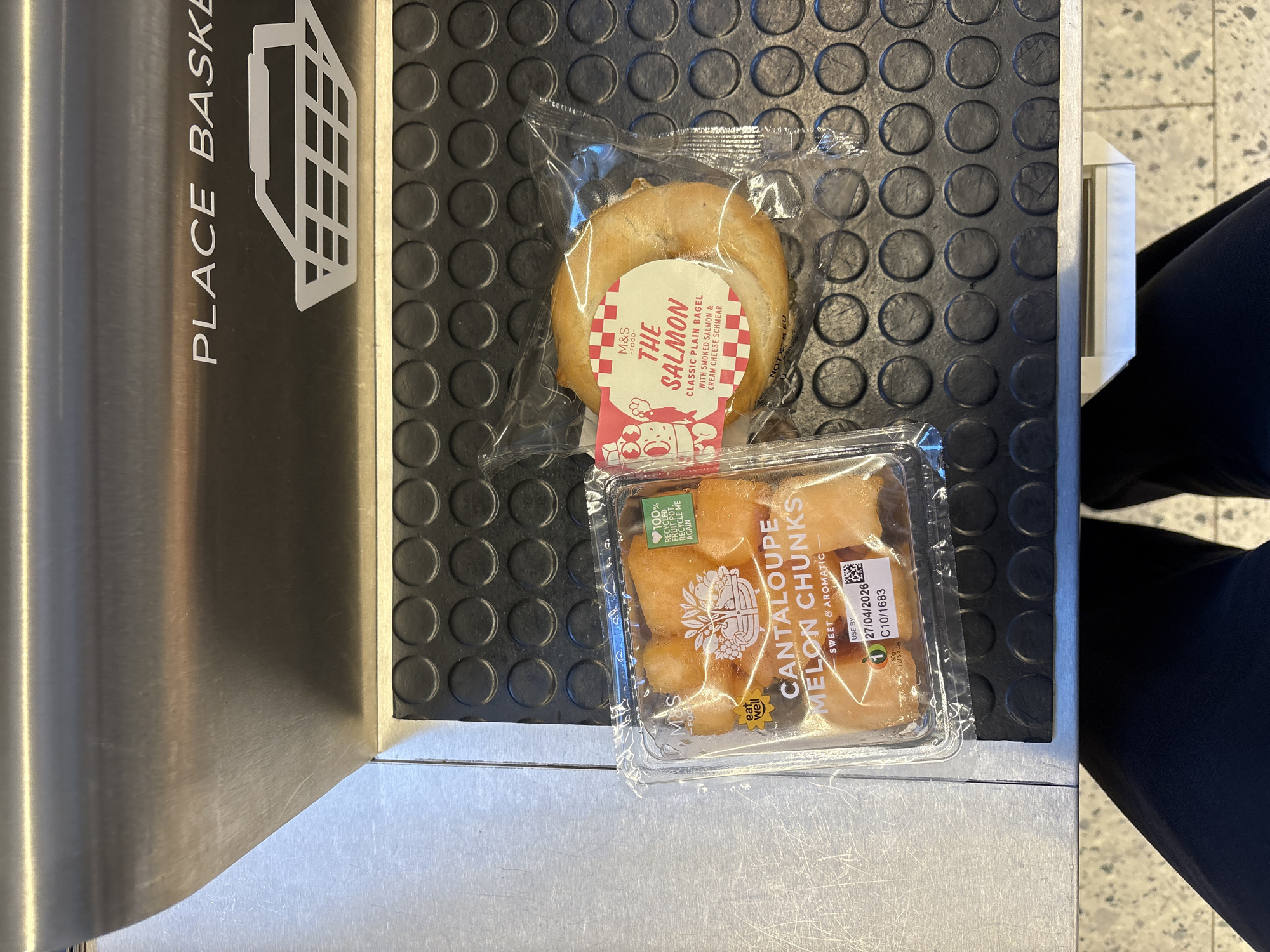 The image captures a close-up shot of a conveyor belt at a supermarket checkout. On the belt are two packaged food items: a salmon bagel from Marks & Spencer (M&S) and a container of cantaloupe melon chunks, also from M&S. A sign indicating "PLACE BASKET" is visible on the side of the conveyor belt. The black surface of the conveyor belt has a patterned texture with circular indentations. A person wearing black trousers is partially visible to the right of the frame. Based on the M&S branding and the overall setting, the location is likely a supermarket in London, United Kingdom.