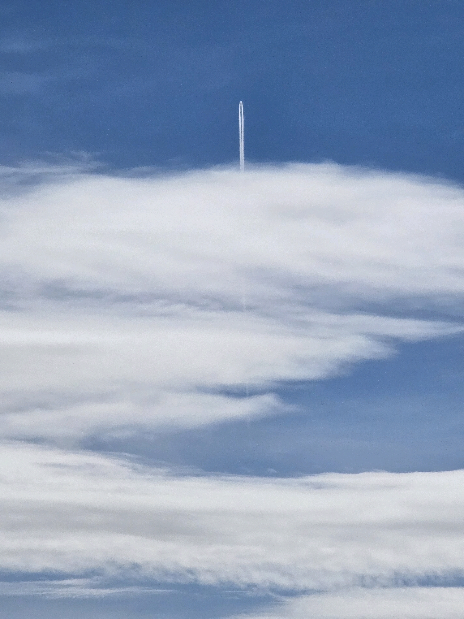 An airplane contrail extends vertically upwards through a blue sky, piercing a layer of fluffy white clouds. The contrail is thin and bright white, with a slightly wider base where it emerges from behind the clouds, and narrows as it ascends into the clear sky. The clouds are dense and opaque in the lower half of the frame, with softer, more wispy formations visible higher up. The overall impression is of a clear day with high-altitude cloud cover, and an aircraft in transit. There is no discernible location in the city of Garmisch-Partenkirchen, Germany, nor any people or specific man-made structures in the image.