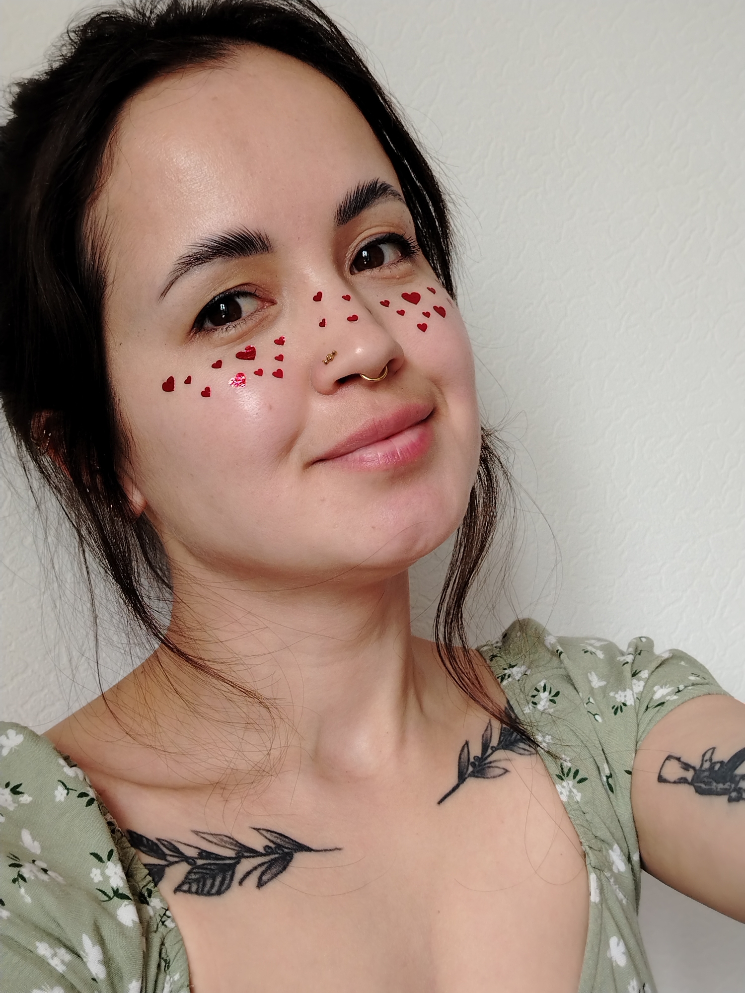 A young woman, likely in Noviy, Russia, is captured in a close-up portrait, looking directly at the camera with a gentle, warm smile. She has dark hair, dark eyes, and well-groomed eyebrows. Her face is adorned with numerous small, red, heart-shaped glitter pieces or stickers scattered across her cheeks, under her eyes, and on her forehead, suggesting a playful or festive mood. She wears gold piercings, including a septum ring and a small stud on her right nostril.
She is dressed in a light green top featuring a white floral pattern. Visible tattoos include a detailed leafy branch design on her left collarbone/chest area and a partially visible abstract or nature-inspired tattoo on her upper right arm. The background is a simple, light-colored wall with a subtle textured finish, indicating an indoor setting. The lighting is bright and even, suggesting daytime conditions. No text is visible in the image.