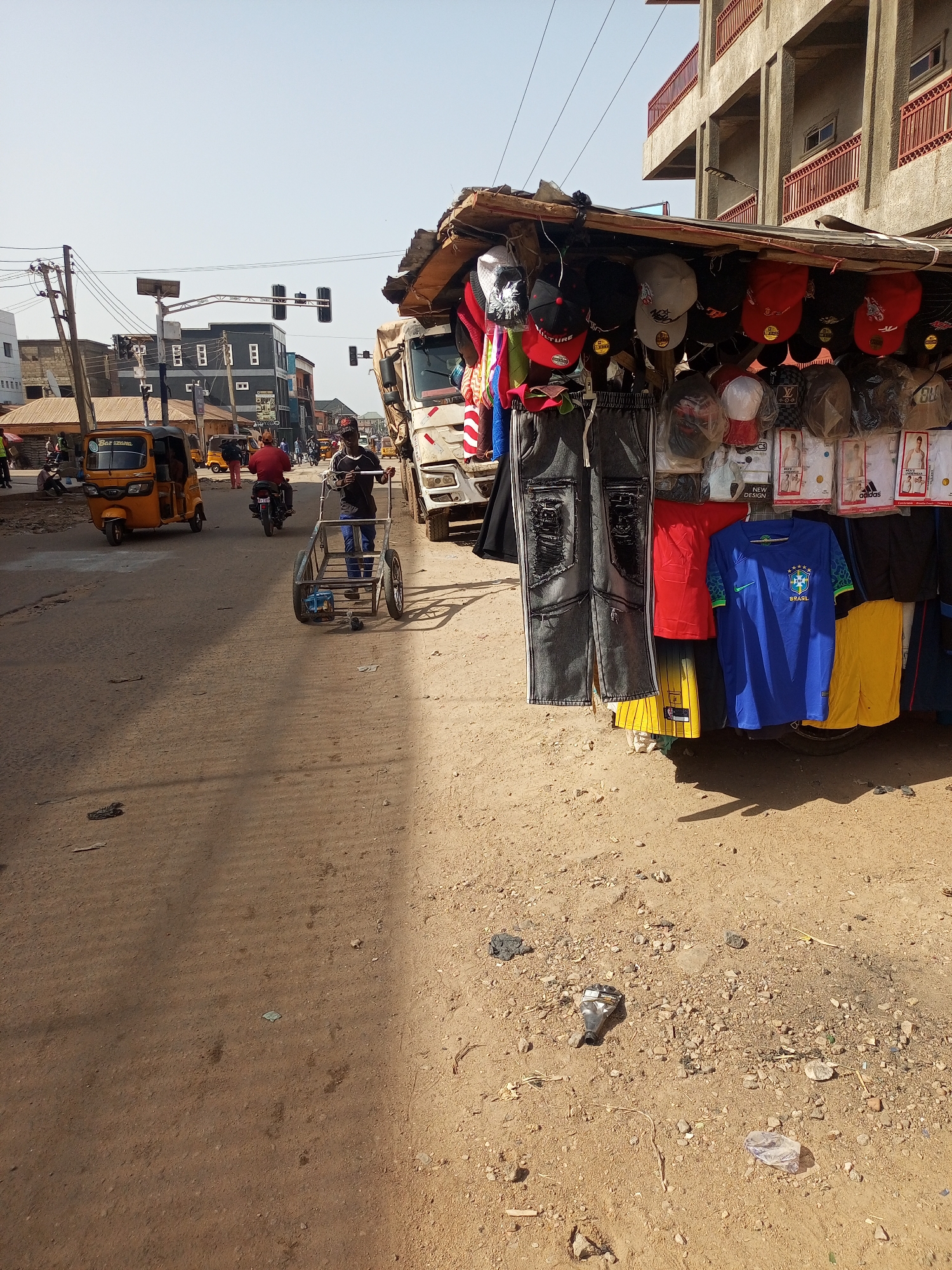 The image depicts a street scene in Maiburiji, Nigeria. The road is unpaved and dusty. A roadside stall displays clothing items, including hats, jeans, shorts, and jerseys. A man is pulling an empty metal cart down the street. There is a tricycle and a motorcyclist behind him. A white truck is parked near the stall. Traffic lights are visible in the distance. Buildings are on the other side of the street. The sky is clear.