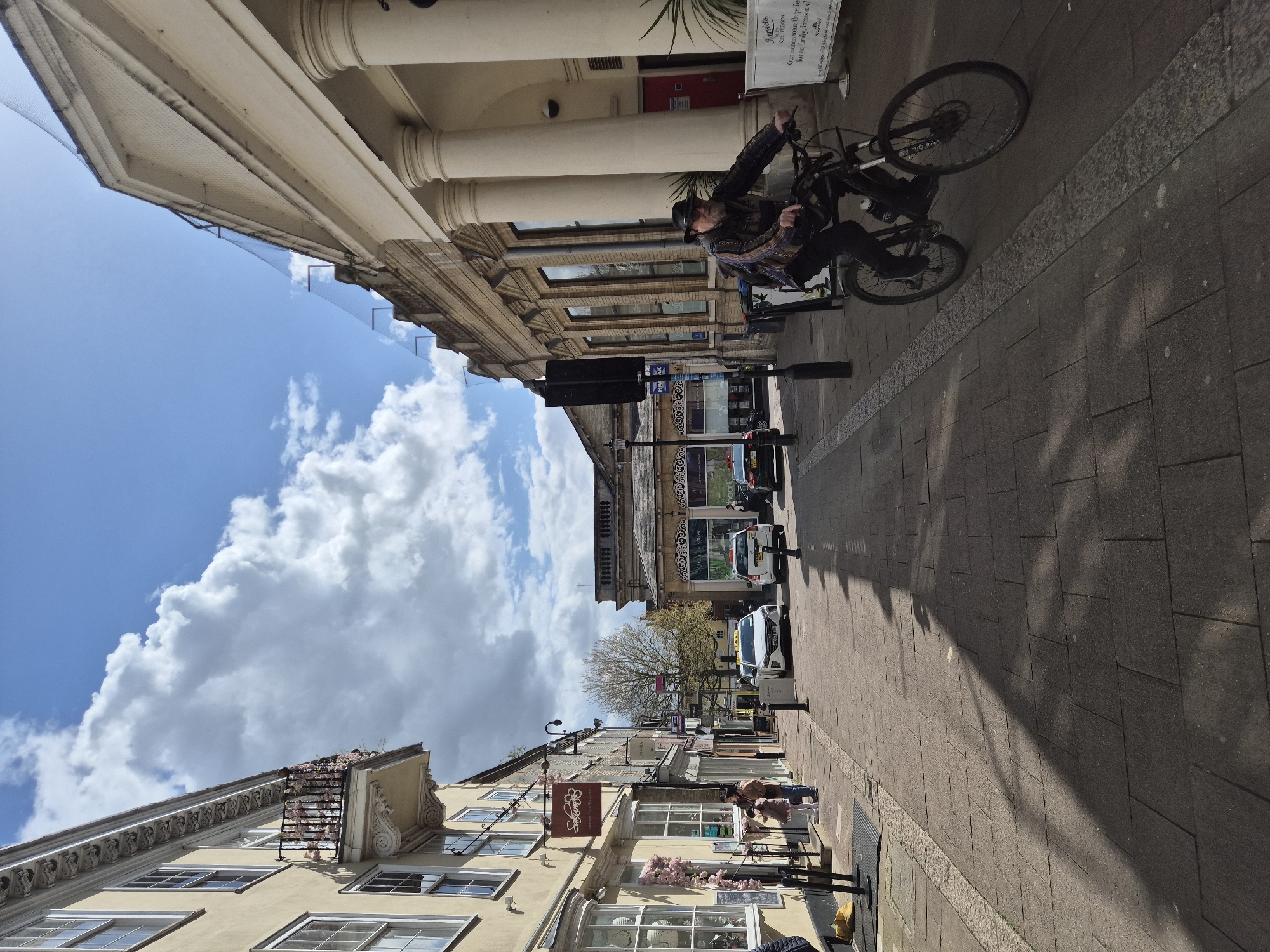 The image captures a street scene in West Suffolk, United Kingdom, under a partly cloudy sky. A cyclist, wearing a hat and jacket, rides his bicycle on the right side of the paved sidewalk, next to buildings. One building features classical architectural elements, including columns. Street signs and shop fronts are visible. Further down the street, parked cars and taxis are seen. The buildings have varied architectural styles, with one having window flower boxes. A shadow is projected across the sidewalk. Banners and advertising signs are attached to the storefronts.
