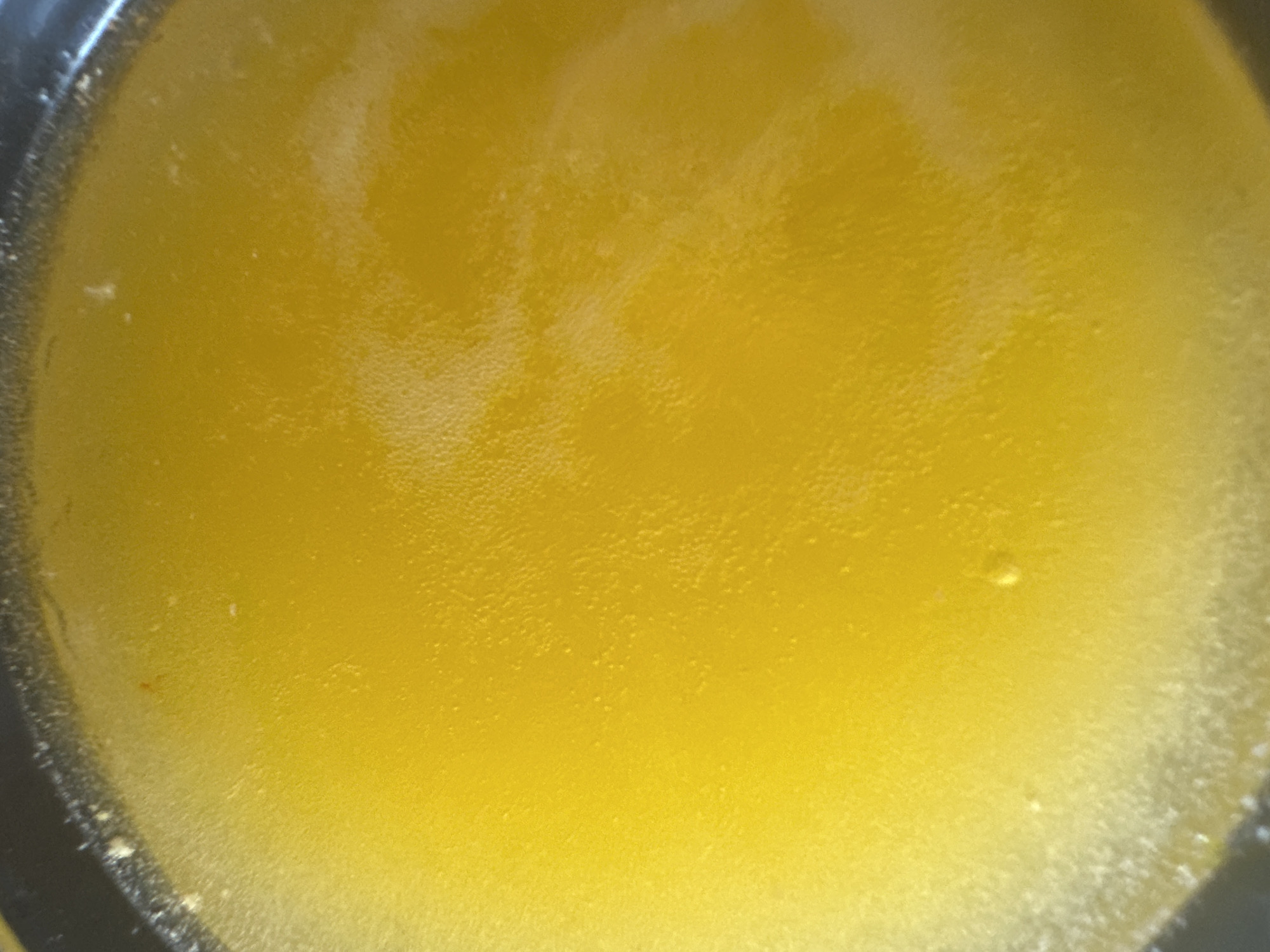 The image is a close-up, overhead view of a shallow, dark-rimmed container filled with a bright yellow, liquid substance. The surface of the liquid appears slightly textured, with a scattering of small bubbles and some lighter, cloudy areas, particularly towards the left edge and in streaks across the surface. The dark rim of the container is visible along the top and left edges of the frame. There are no people or distinct objects within the liquid itself, only the texture and sheen of the substance. The location in Novoaltaysk, Russia, cannot be determined from the visual information provided in the image.