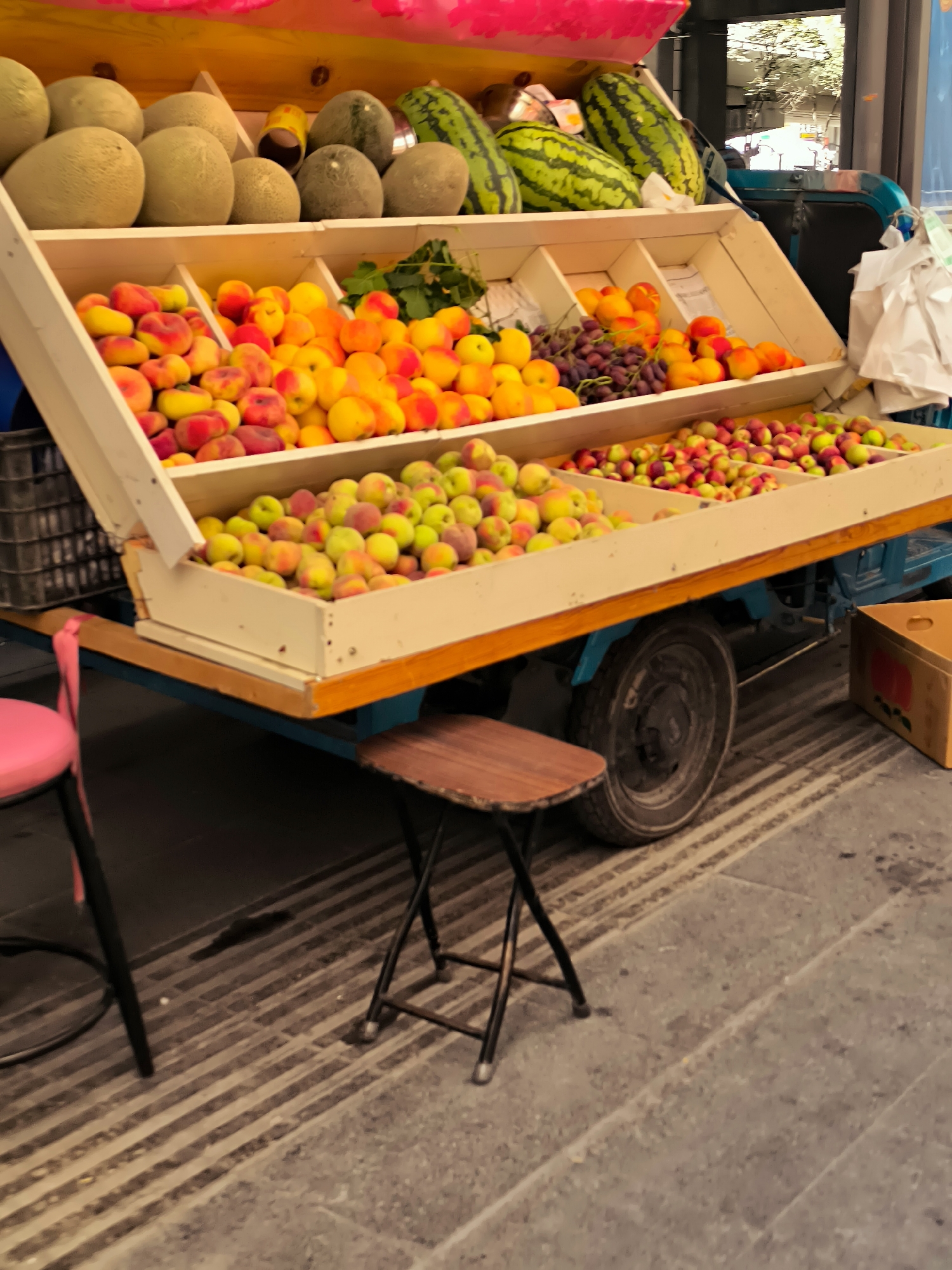 A street market stall in Ürümqi, China, displays a variety of fresh fruits. The stall is a multi-tiered cart with a yellow and pink canopy. The top tier holds several large, round melons, some striped like watermelons and others appearing to be cantaloupes or honeydew melons. The second tier is filled with ripe peaches and apricots, a vibrant mix of reds, oranges, and yellows. Below that, a large tray overflows with smaller fruits, possibly plums or nectarines, also in various shades of red, green, and yellow. A bunch of dark purple grapes is also visible on the second tier, nestled amongst the peaches.
The cart has a blue frame and a wooden base. Two small stools are positioned in front of the cart: a pink stool on the left and a brown wooden stool on the right. In the background, a portion of another vehicle and some white plastic sheeting are visible, along with the blurred suggestion of buildings and trees, indicating an urban setting. The ground is paved with alternating light gray and dark gray striped tiles. A cardboard box sits to the right of the cart. The overall scene suggests a bustling marketplace scene.